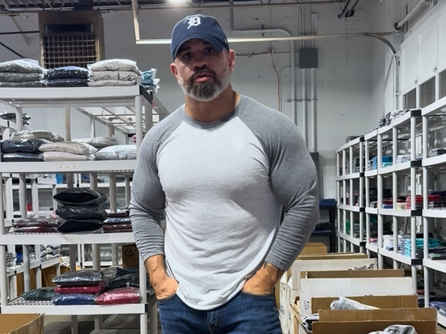 Man wearing a cap and gray shirt standing in a warehouse setting with shelves and boxes in the background.