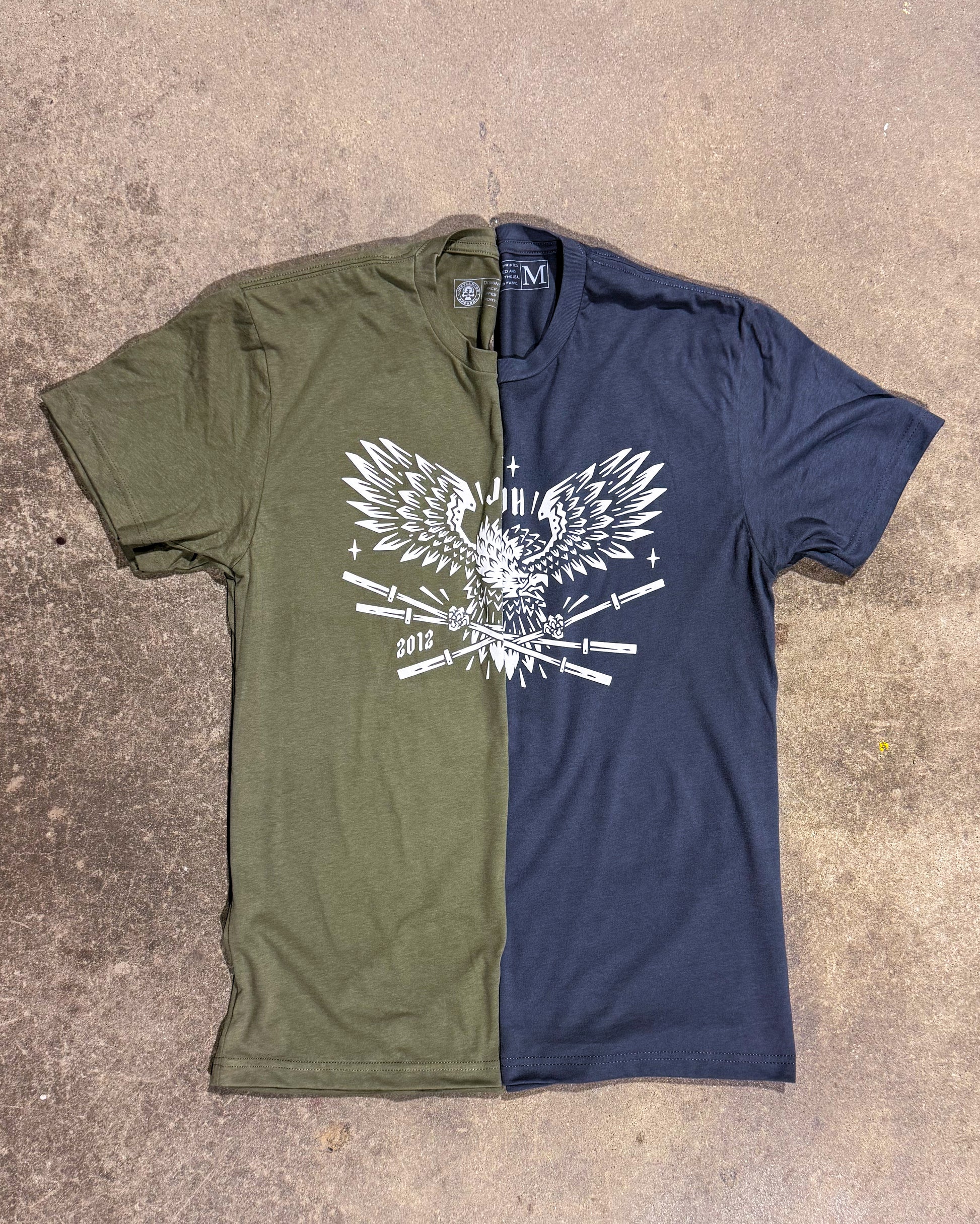 Two tshirts folded in half on cement to make one tshirt. One OD green, the other grey both with an eagle holding barbells on it. 