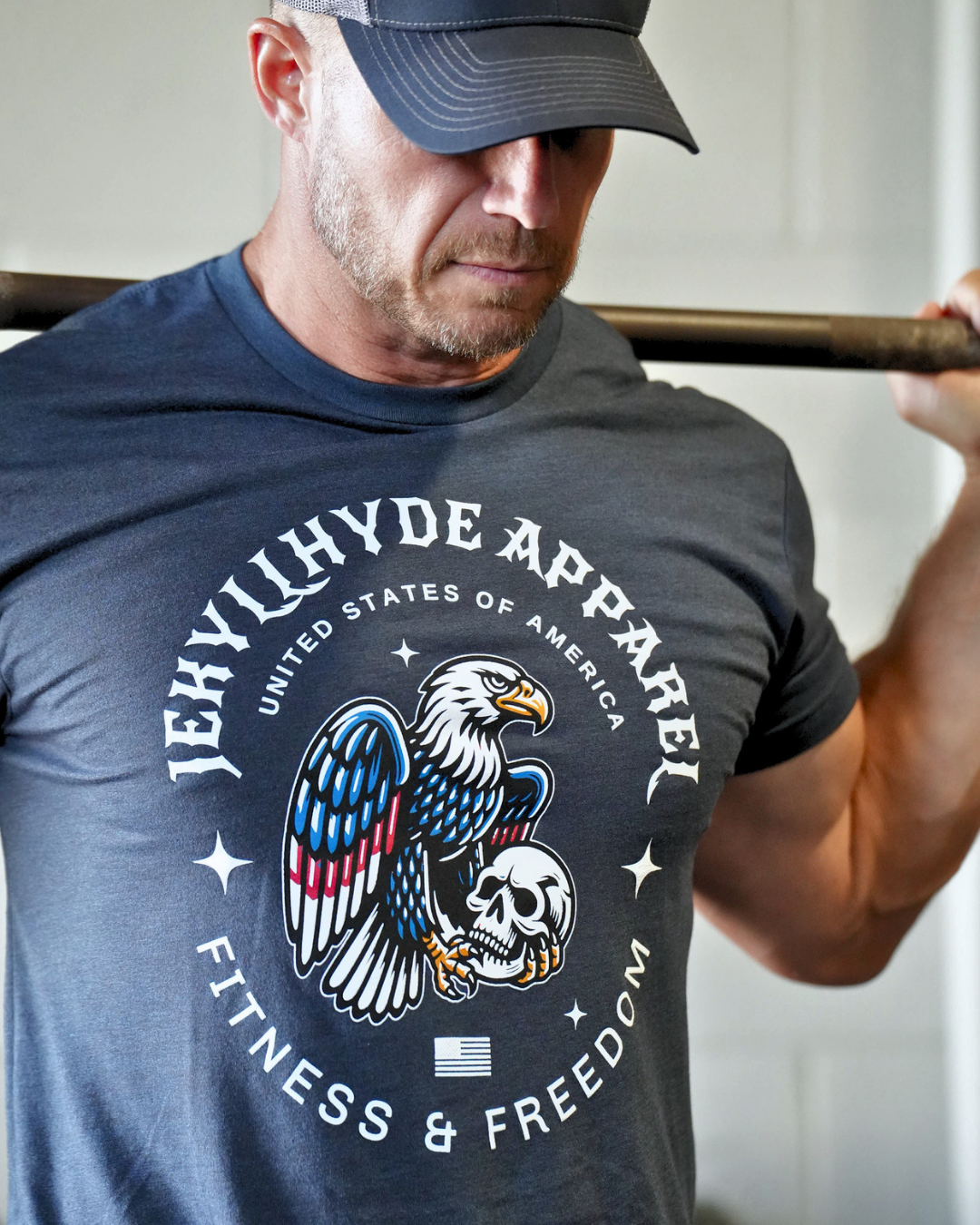 A man wearing a dark NAVY t-shirt with a large graphic of an eagle and skull, and a baseball cap, lifting a barbell. The t-shirt features the text 'JEKYLHYDE APPAREL' and 'UNITED STATES OF AMERICA FITNESS & FREEDOM'.