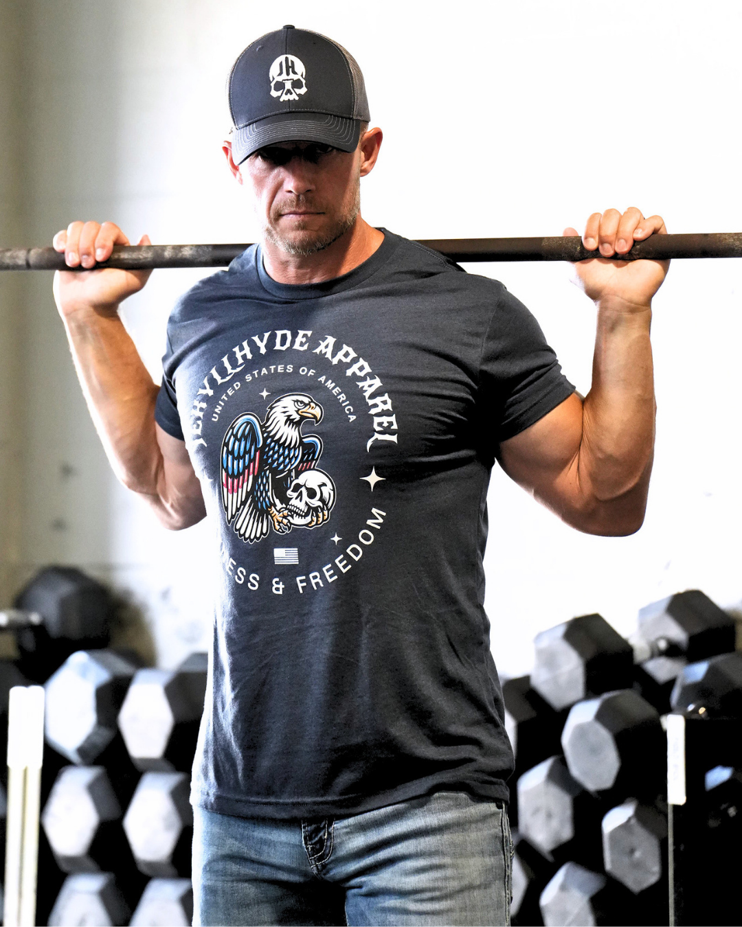 A man in a gym wearing a black t-shirt with a graphic of an eagle and skull and the text 'ANTHILYHIDE APPAREL' and 'fitness & FREEDOM', holding a barbell on his shoulders.