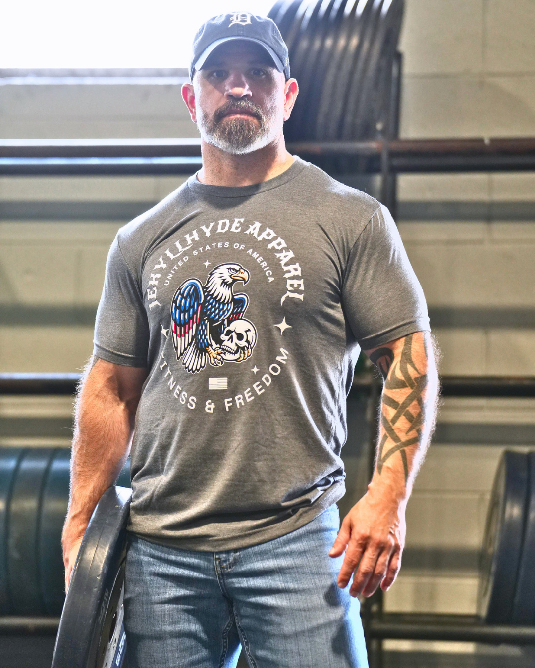 A man in a gym wearing a heather gray t-shirt with a patriotic eagle graphic and the text 'JEKYLLHYDE APPAREL' and 'FINTESS AND FREEDOM'.
