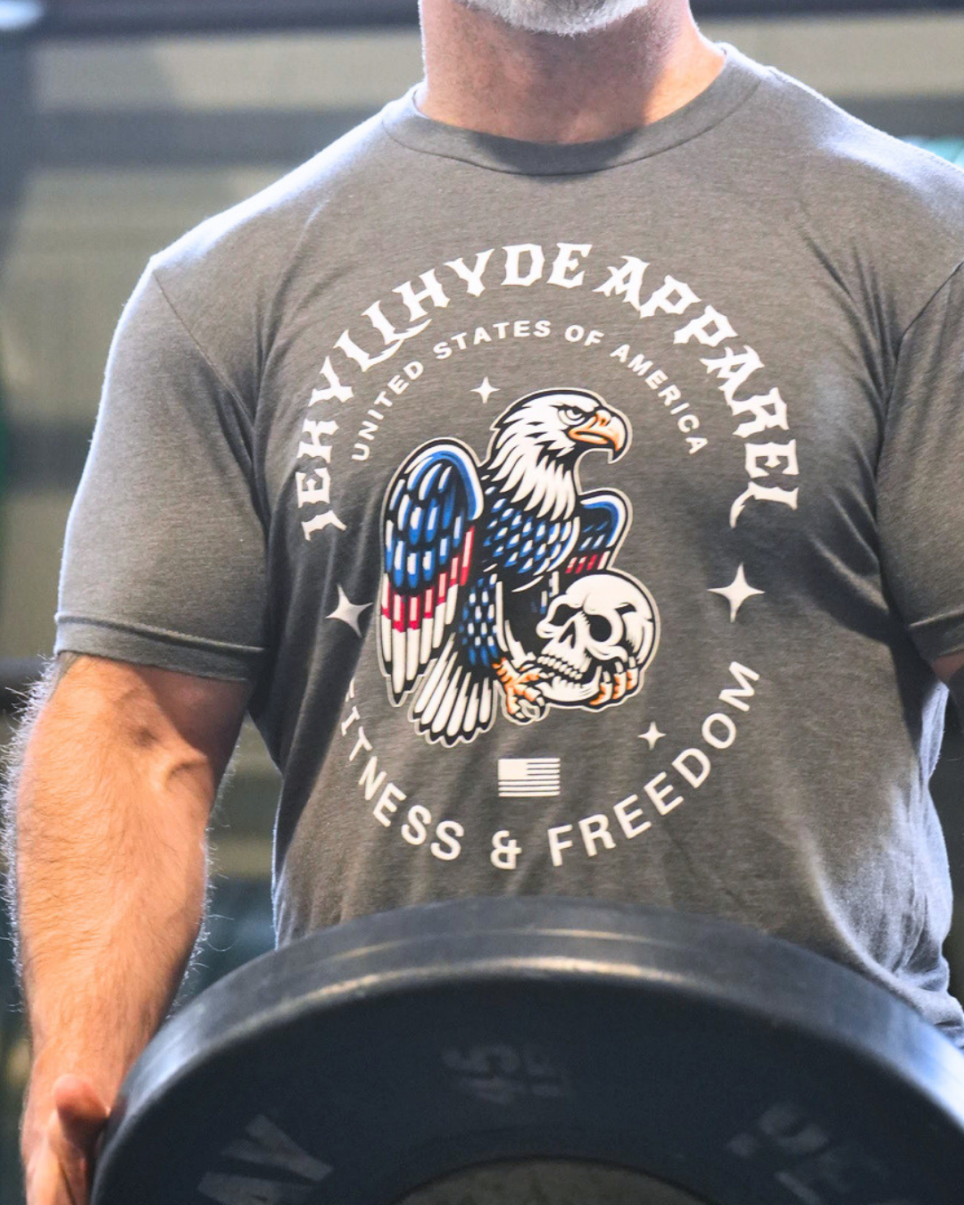 A close-up of a man wearing a heather gray t-shirt with a large graphic print featuring an eagle, skull, and the text 'Jekyll Hyde Apparel' and 'Fitness & Freedom'. He is holding a barbell in a gym setting.