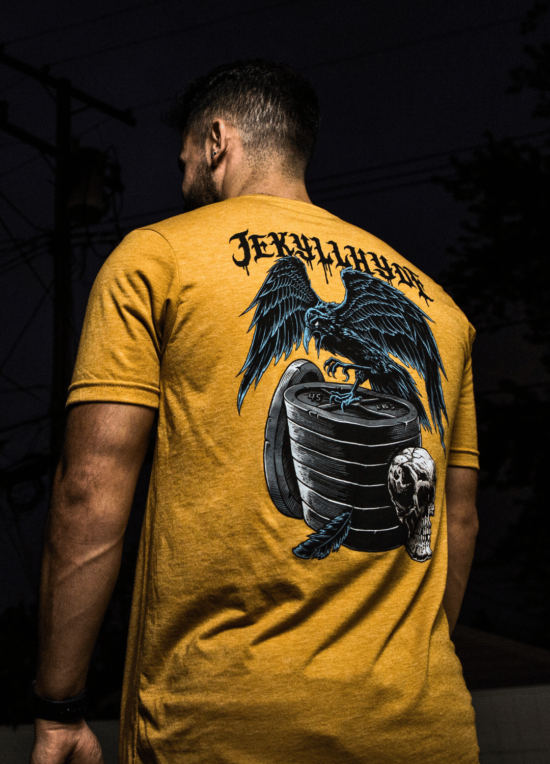 Man standing with his back to the camera wearing a gold Raven and skull tshirt. 
