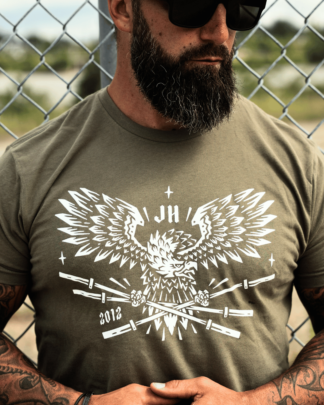 A man standing against a fence wearing a OD green t-shirt with a bald eagle holding barbells in it's claws. 