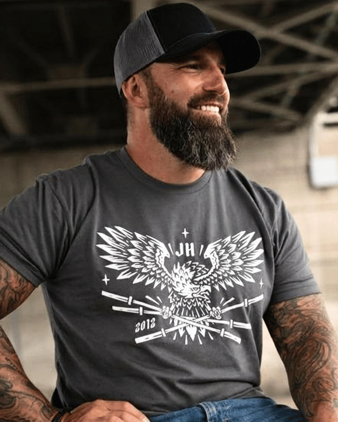A man standing sitting outside wearing a grey t-shirt with a bald eagle holding barbells in it's claws. 