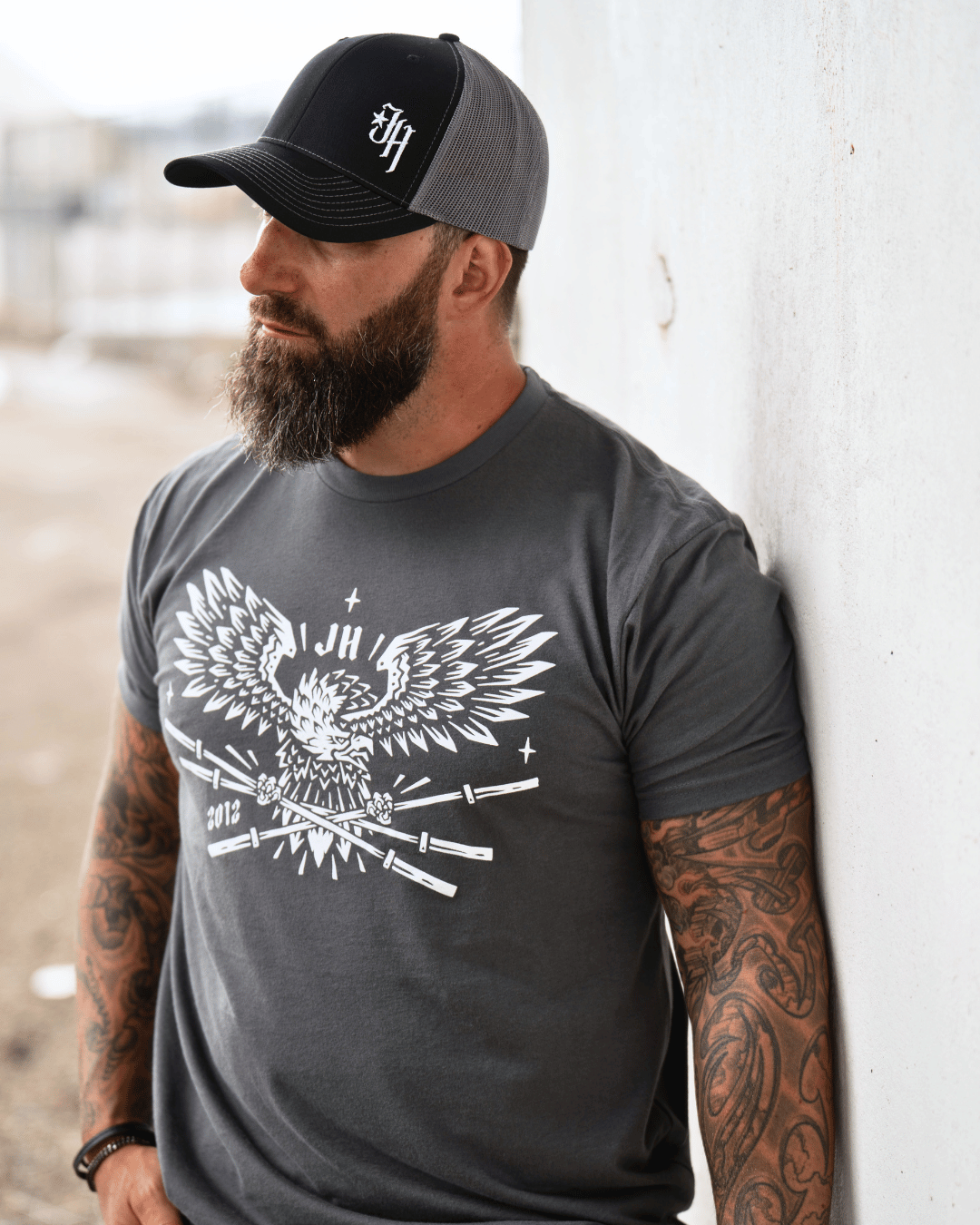 A man standing against a wall wearing a grey t-shirt with a bald eagle holding barbells in it's claws. 