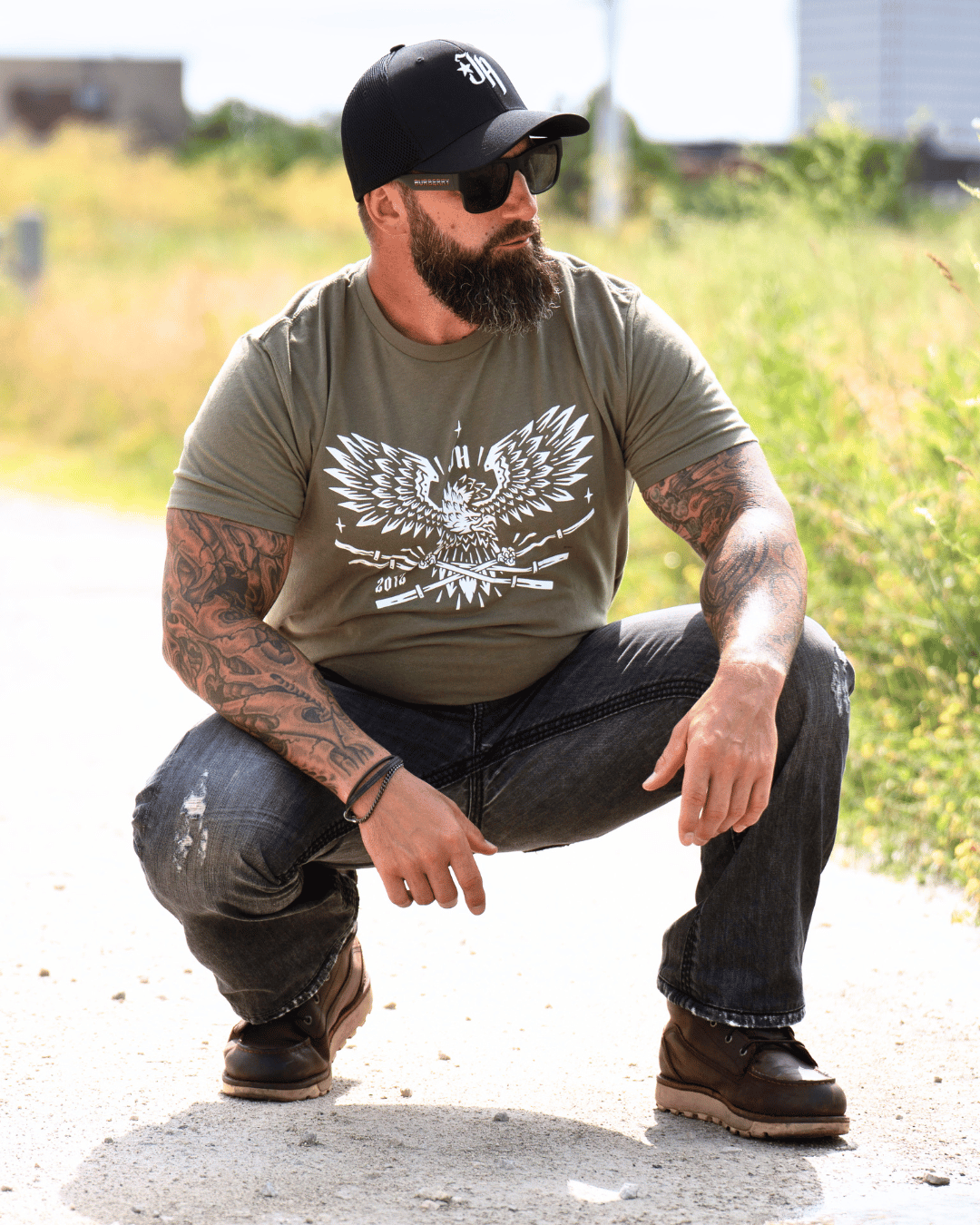 A man crouching down wearing a OD green t-shirt with a bald eagle holding barbells in it's claws. 