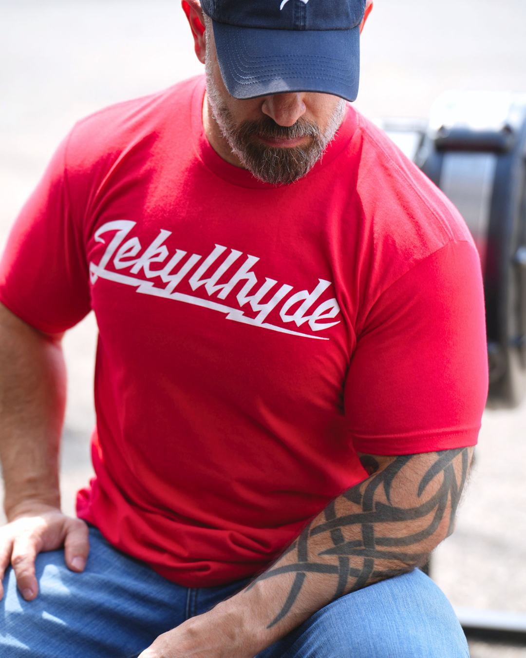A man wearing a red crewneck t-shirt with 'Jekyllhyde' printed in white script across the chest, paired with blue jeans and a navy blue baseball cap.