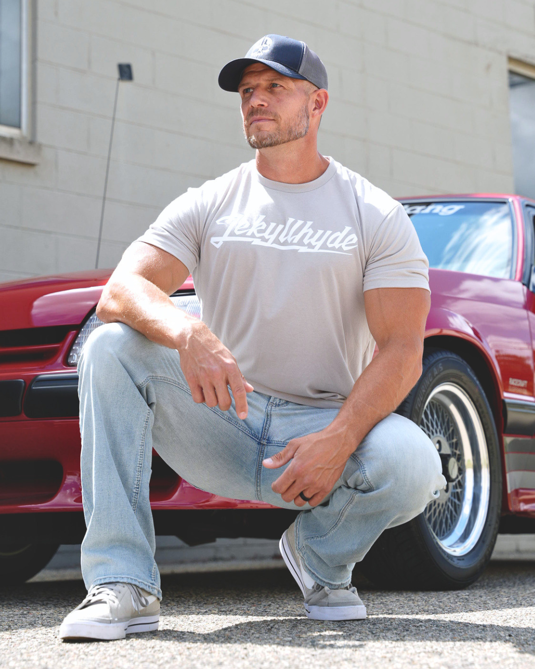 A muscular man wearing a light gray t-shirt with 'Jekyllhyde' printed across the chest, light blue jeans, and a dark baseball cap crouches in front of a red classic car.