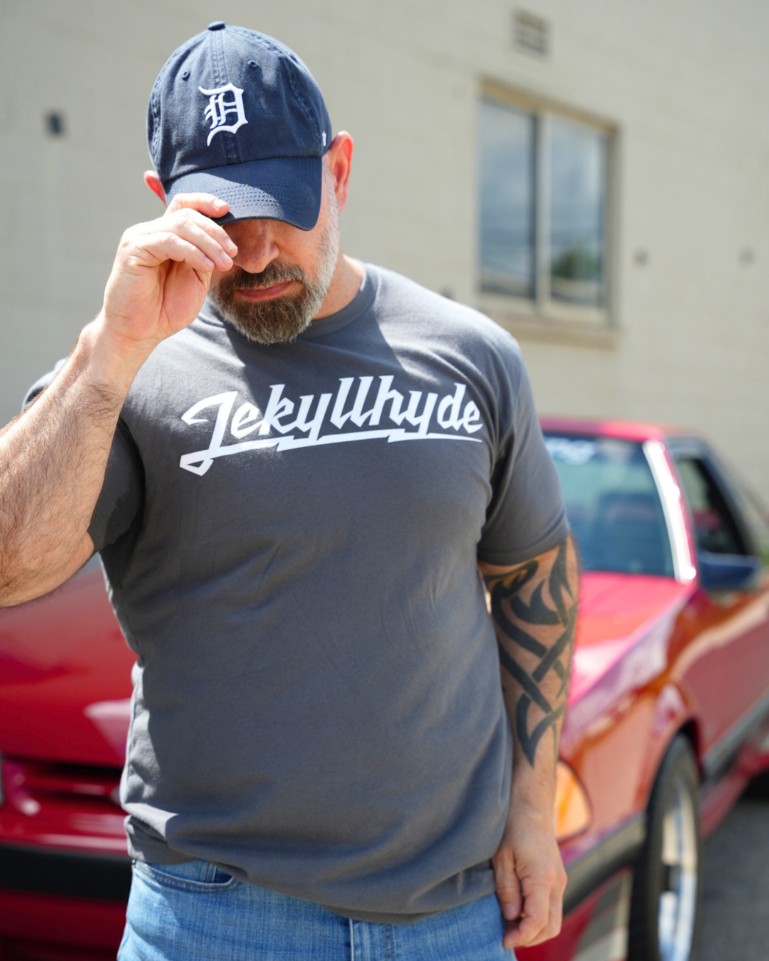 A man wearing a dark gray t-shirt with 'Jekyllhyde' printed in white script across the chest, paired with a navy blue baseball cap and blue jeans, standing outdoors in front of a red car.