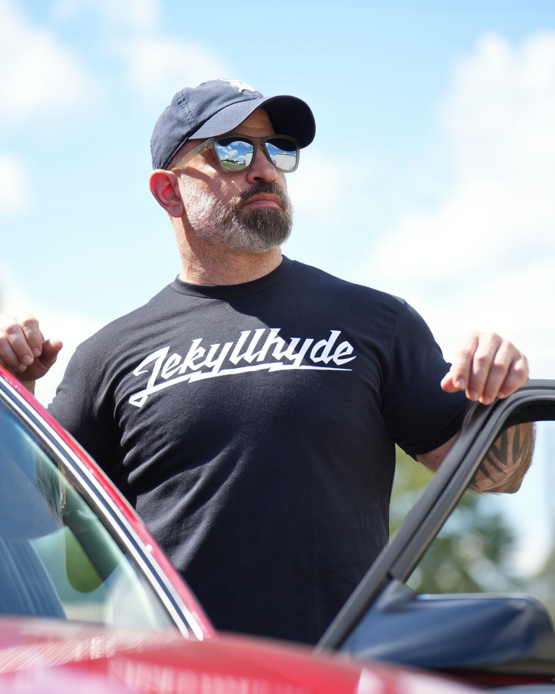 A man wearing a black crewneck t-shirt with 'Jekyllhyde' printed in white script across the chest, standing next to a red car outdoors under a blue sky.