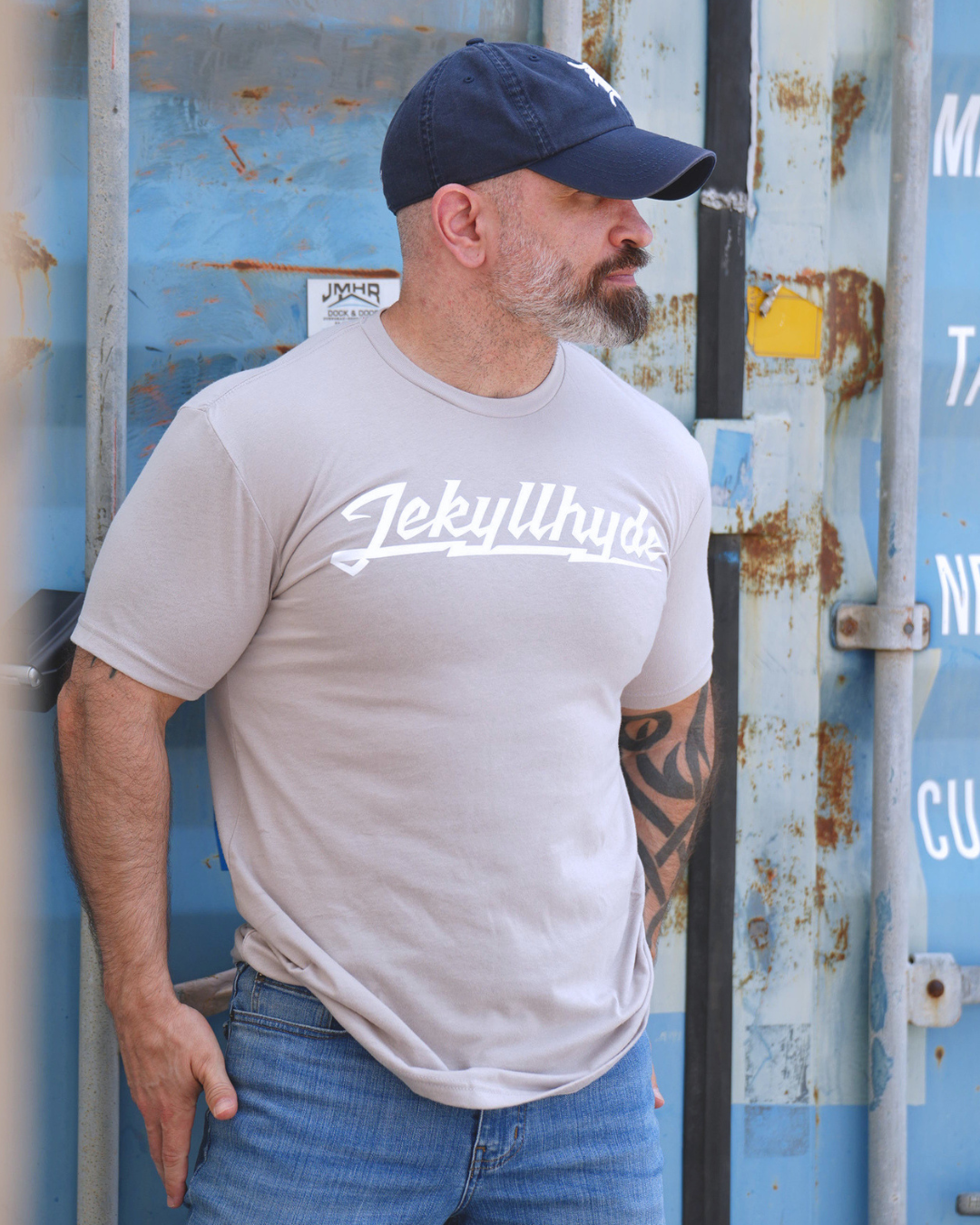 A man wearing a light grey crewneck t-shirt with 'JekyllHyde' printed in white script across the chest, paired with a navy blue baseball cap and blue jeans, standing in front of a weathered blue shipping container.