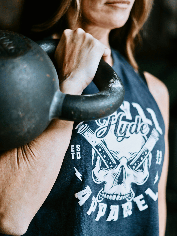 Woman with a kettlebell on her shoulder wearing a blue skull crop tank. 