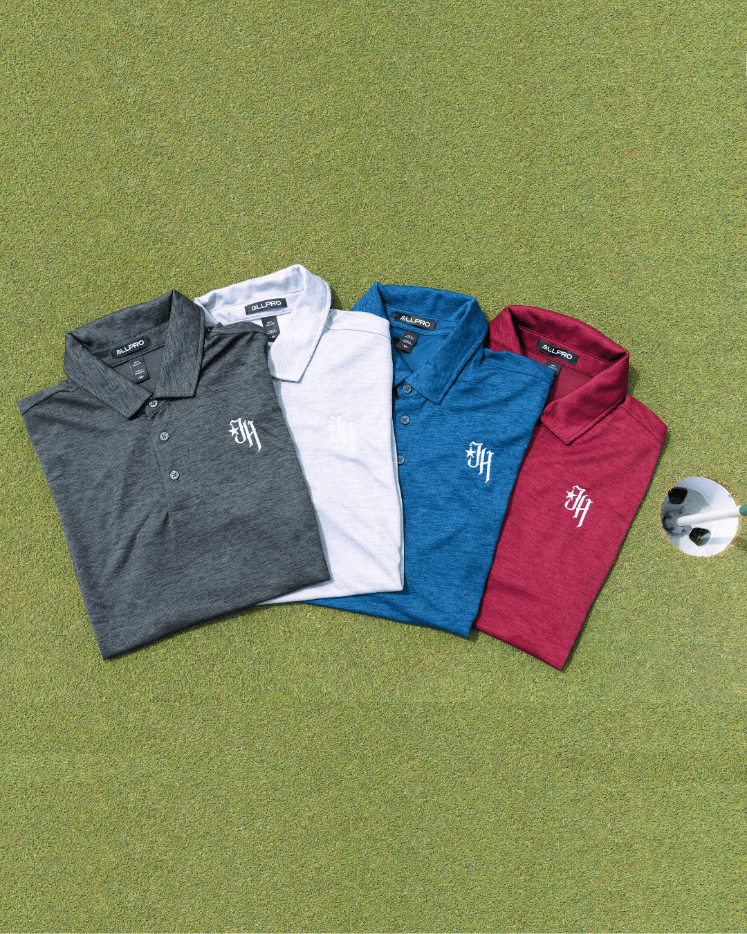 Four folded polo shirts in gray, white, blue, and red on a green surface with a golf ball.