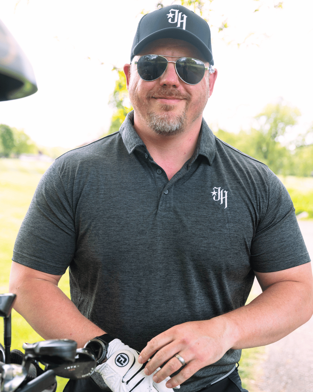 Man wearing a dark gray polo shirt with a logo, black cap, and sunglasses outdoors.