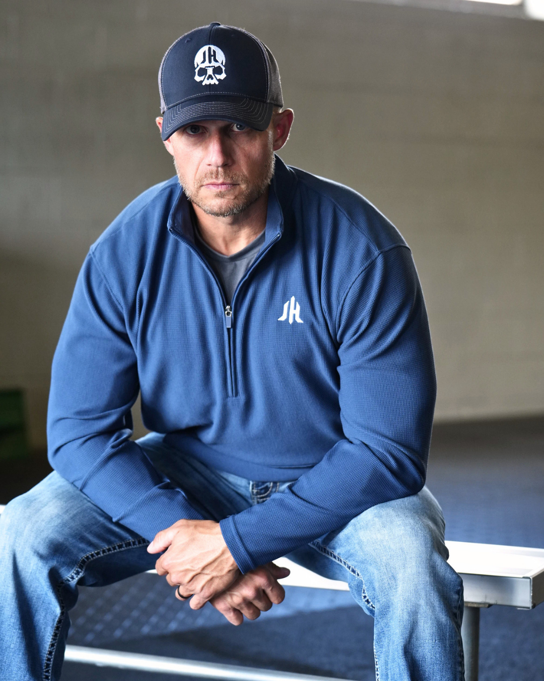 A man wearing a navy blue quarter-zip pullover with a white embroidered logo on the left chest, blue jeans, and a dark baseball cap with a white skull graphic, sitting on a bench in a concrete structure.