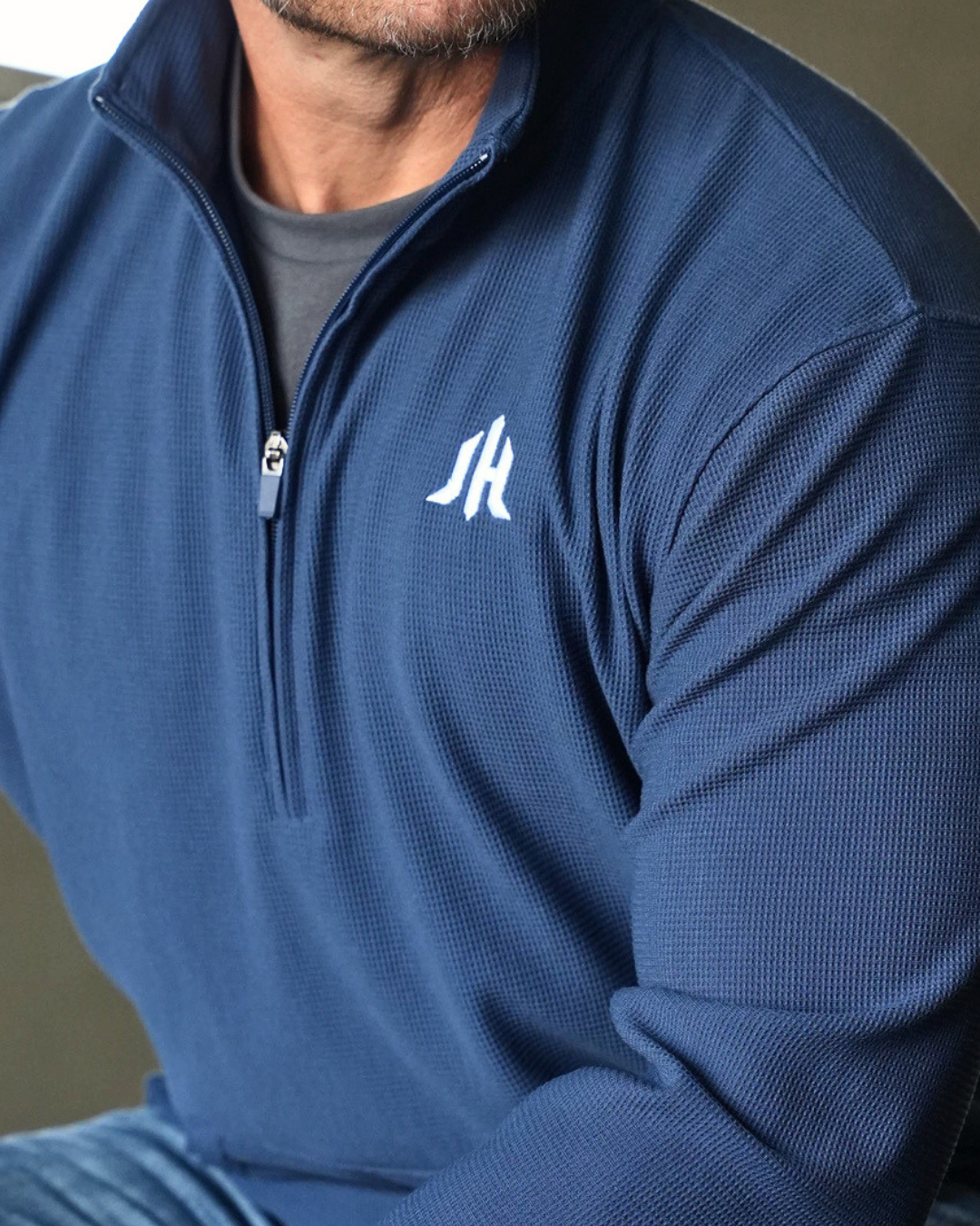 A close-up of a man wearing a navy blue waffle-knit quarter-zip pullover with a white stylized 'JH' logo on the left chest, layered over a gray t-shirt.