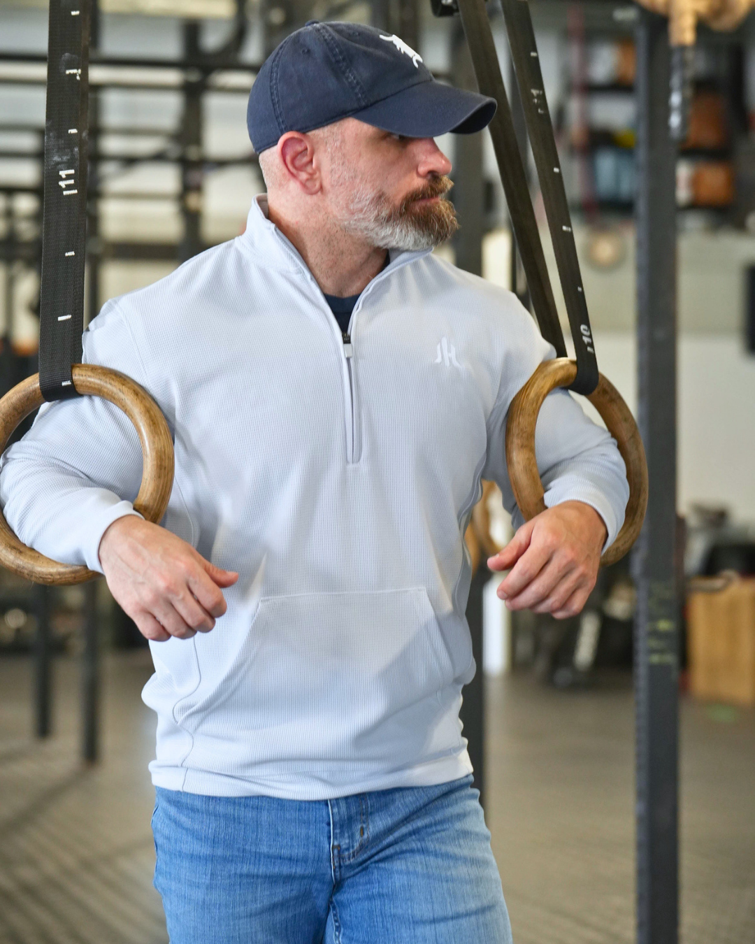 A man in a gym wearing a light blue quarter-zip pullover and blue jeans, standing in front of gymnastic rings.