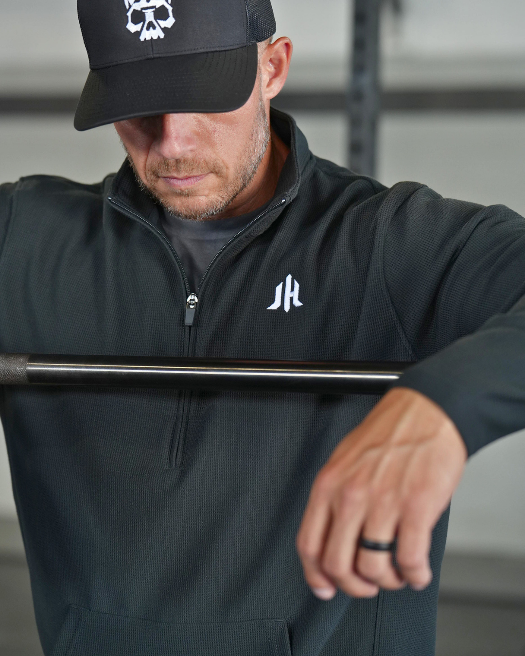 A man wearing a black quarter-zip pullover with a white stylized 'JH' logo on the left chest, paired with a black baseball cap featuring a white skull graphic. He is holding a barbell in a gym setting.
