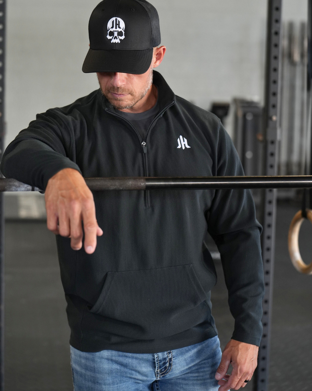 A man in a gym wearing a black quarter-zip pullover with a white logo on the left chest, a black cap with a skull and 'JH' logo, and blue jeans.