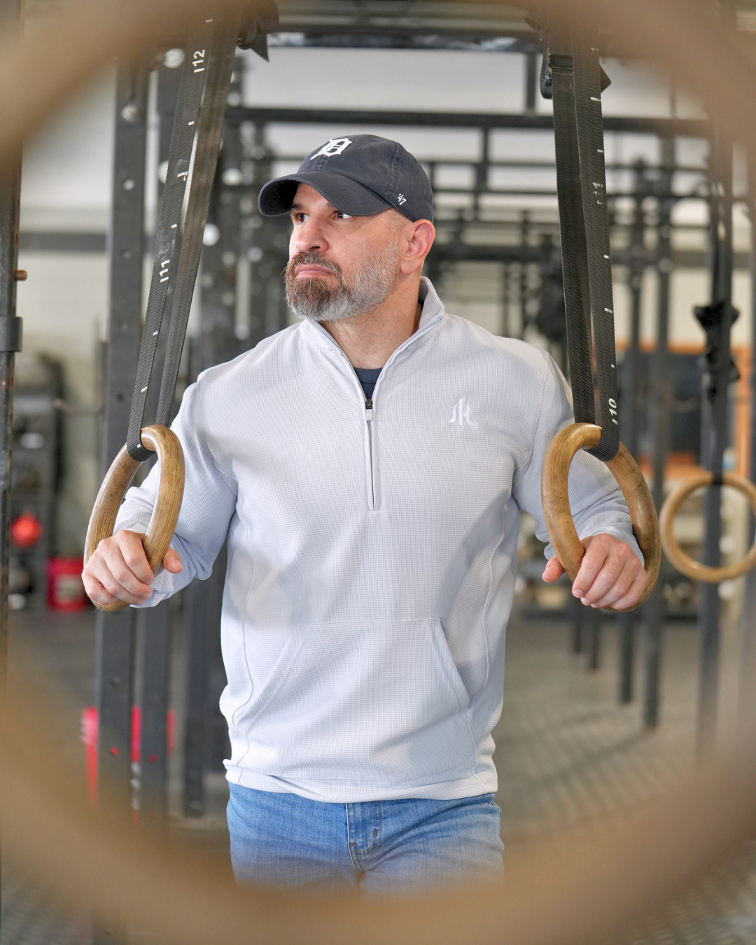 A man in a gym wearing a light gray quarter-zip pullover and blue jeans, holding gymnastic rings. The photo is framed by a blurred circular foreground element.
