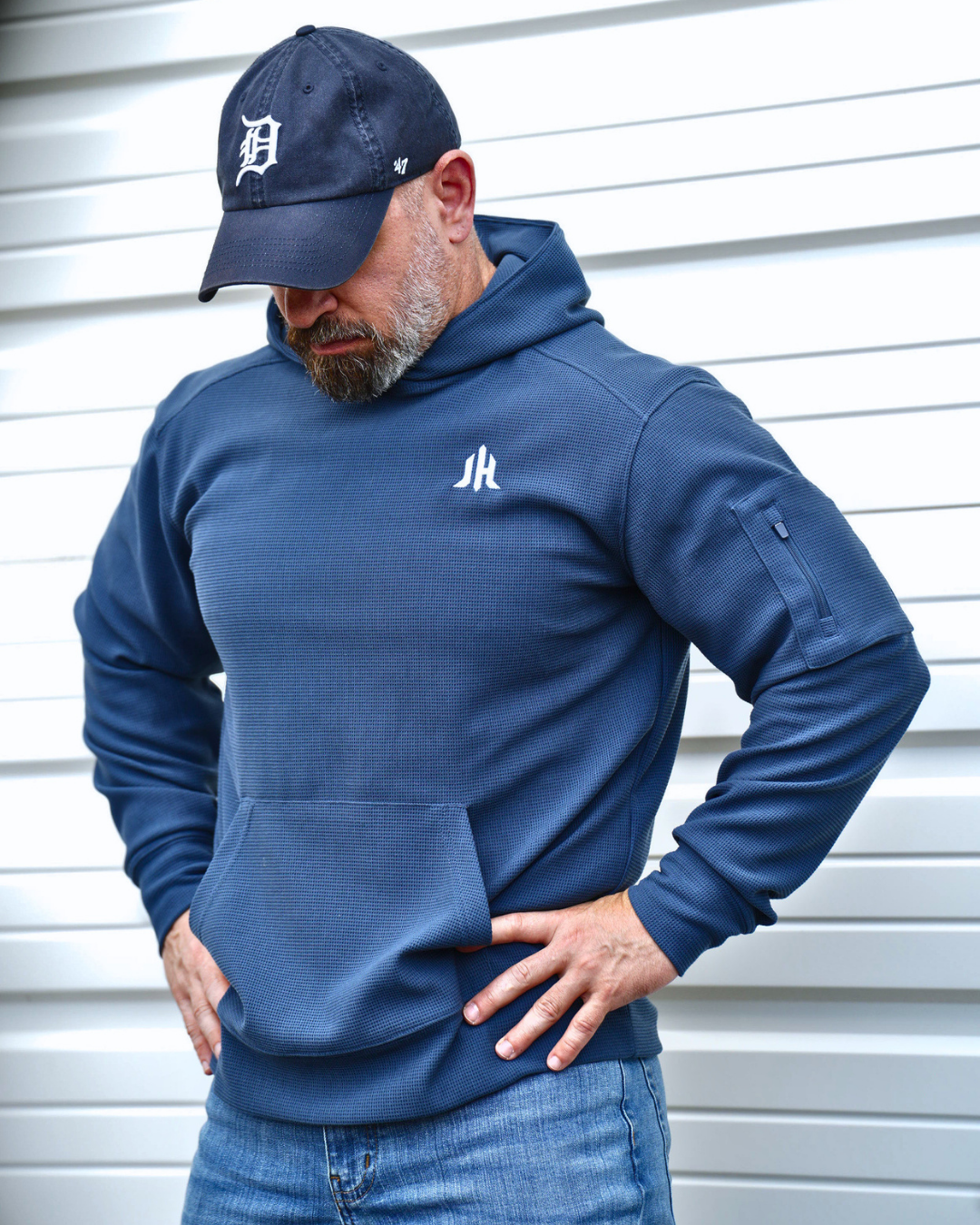 A man wearing a navy blue pullover hoodie with a white embroidered logo on the left chest, paired with a matching navy baseball cap and blue jeans, standing against a white siding background.