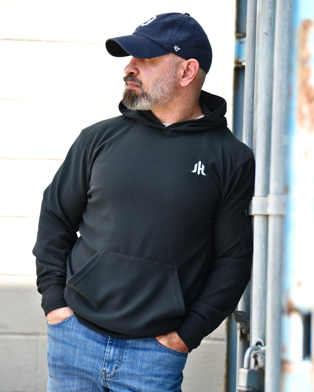A man wearing a black pullover hoodie with a small white stylized logo on the left chest, paired with blue jeans and a dark navy baseball cap, standing outdoors next to a metal structure.
