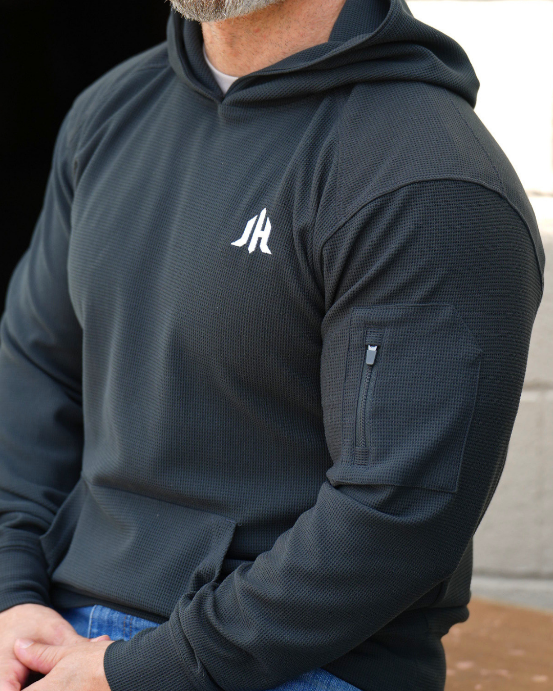 A man wearing a dark charcoal gray hooded sweatshirt with a white stylized 'JH' logo on the left chest and a zippered sleeve pocket, shown in a close-up shot.