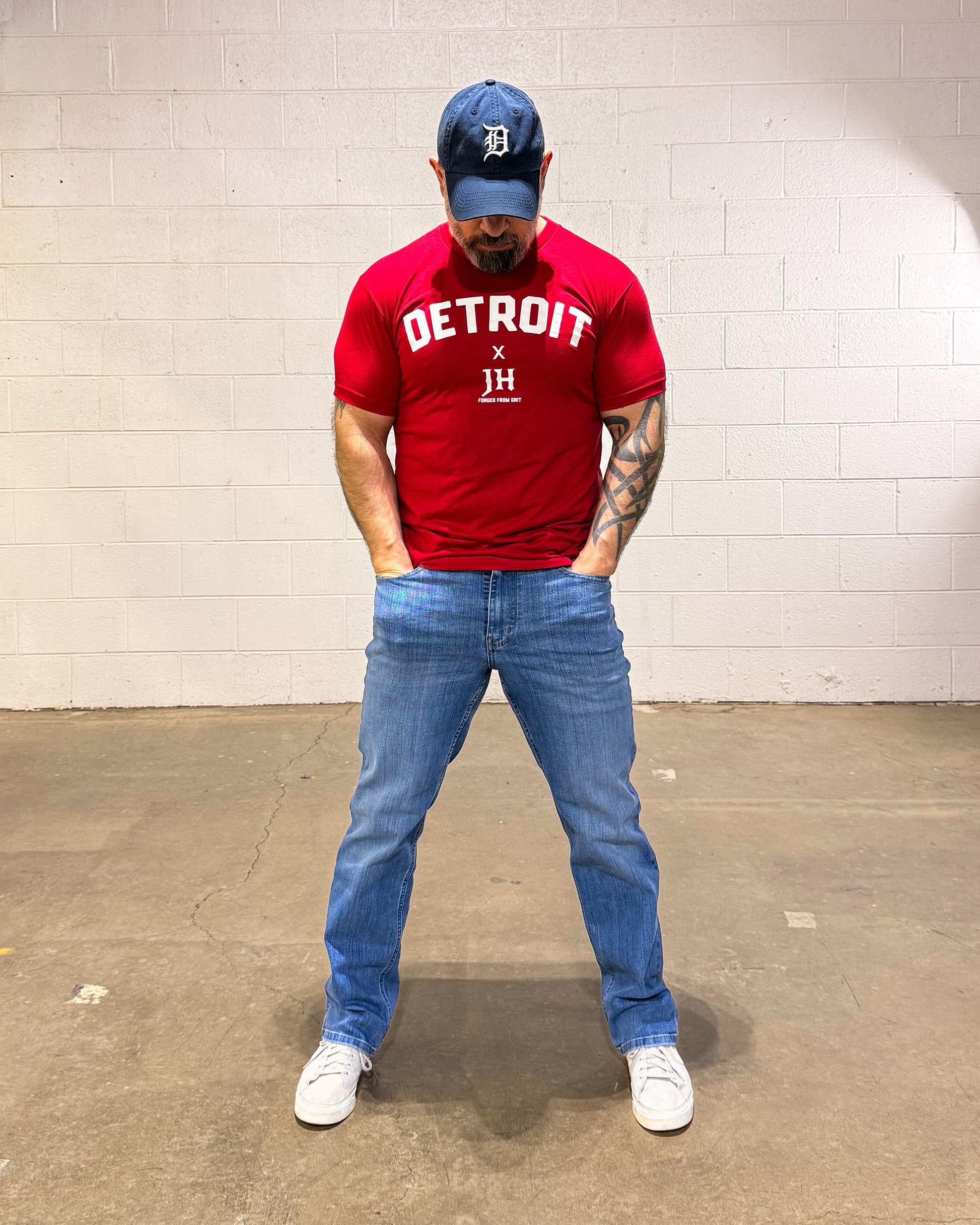 Person wearing a red 'Detroit' t-shirt, blue jeans, and a cap in an indoor setting.