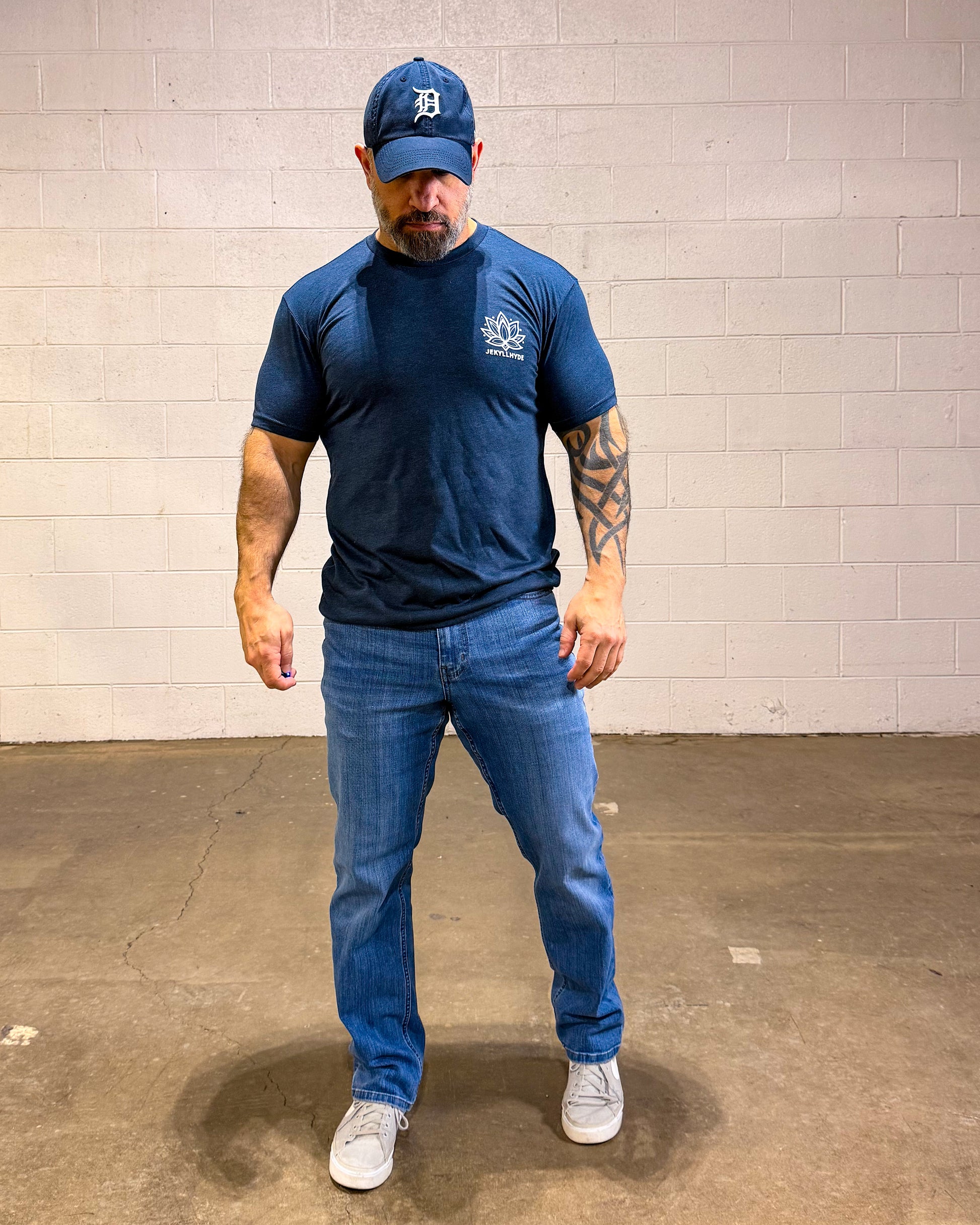Man wearing a navy t-shirt, blue jeans, and a navy cap with a logo against a plain wall.