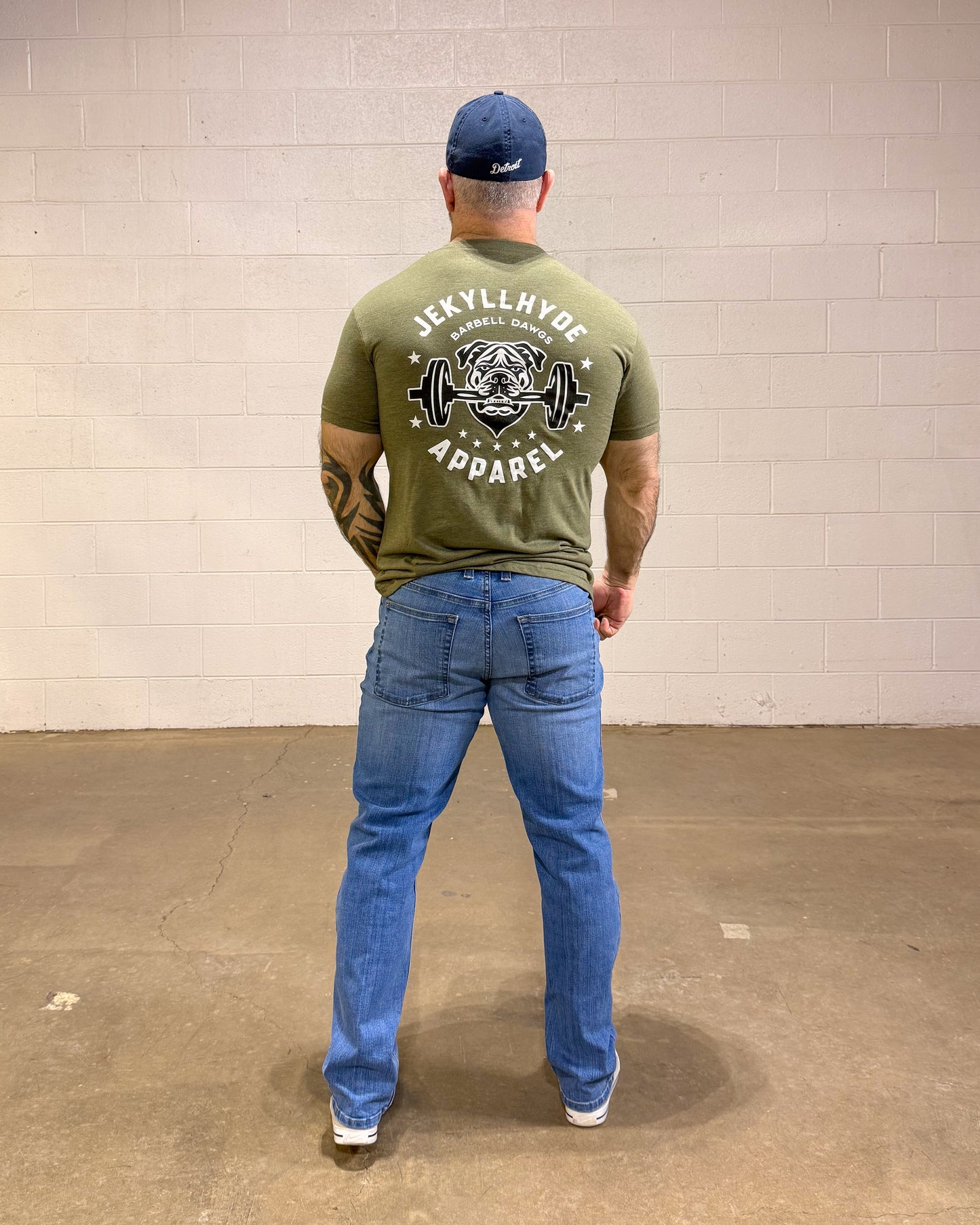 Person wearing a green t-shirt with branding and blue jeans standing against a plain wall.
