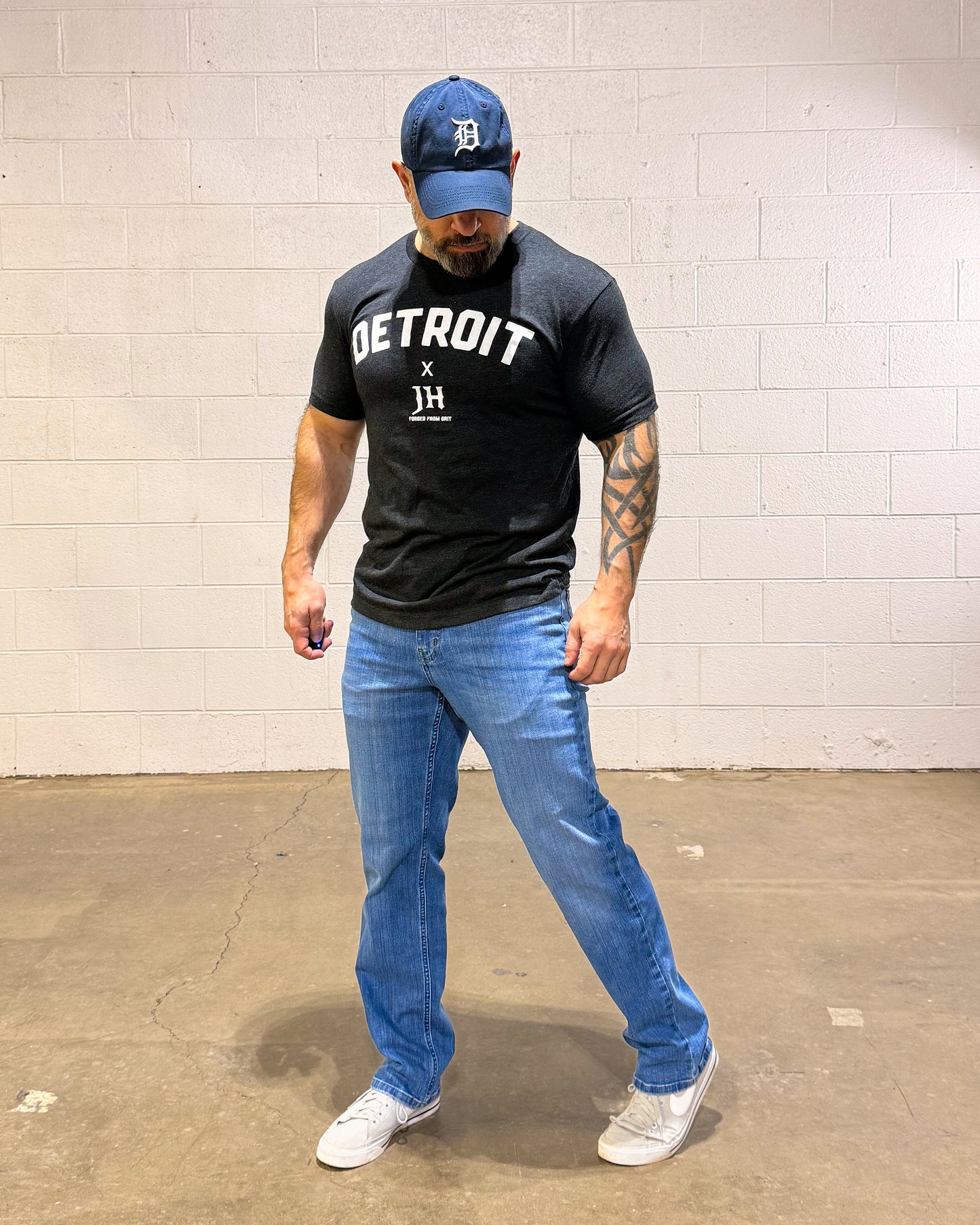 Man wearing a black 'Detroit' t-shirt, blue jeans, and a blue cap against a white brick wall.