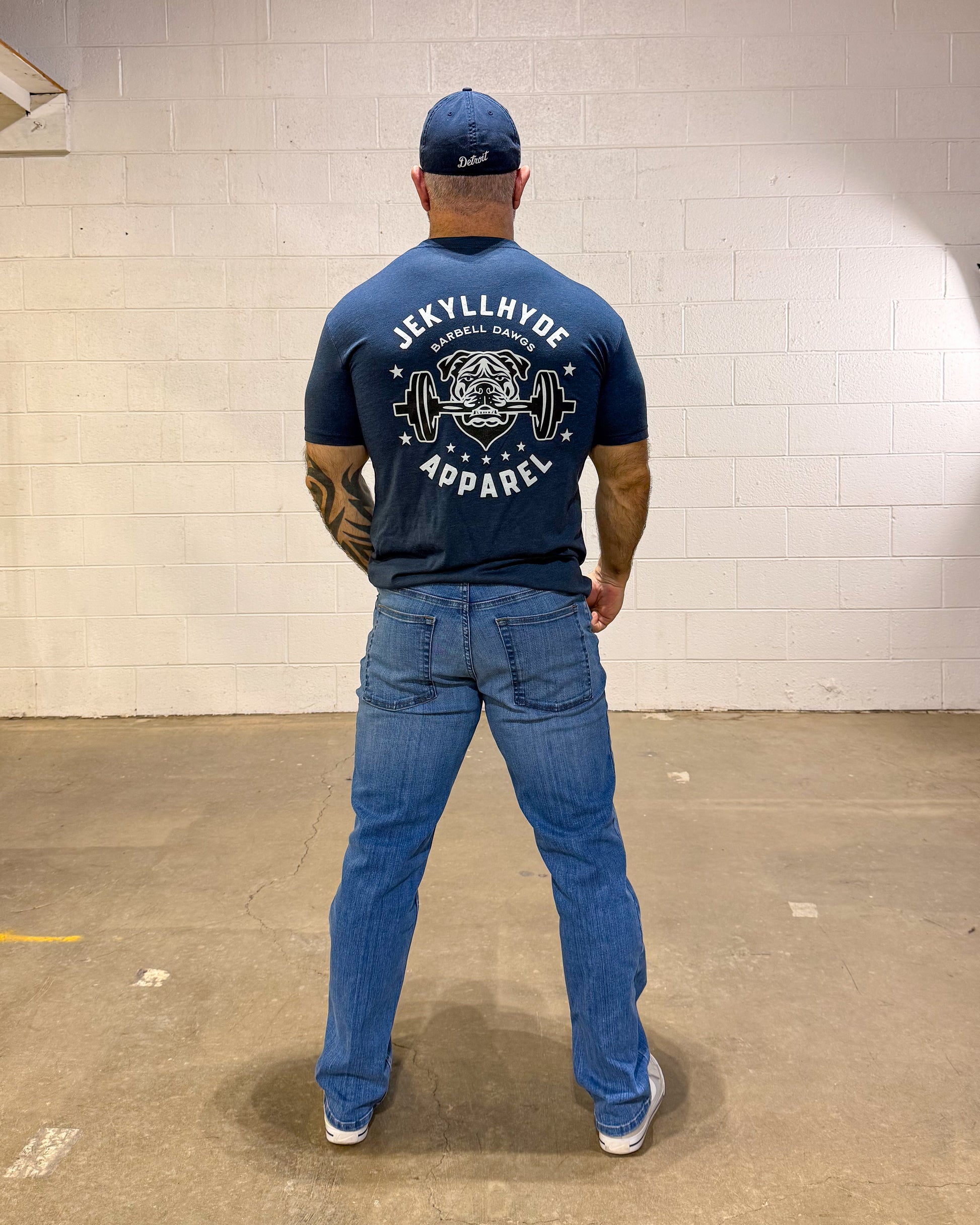 Person wearing a blue t-shirt and jeans with a visible brand logo, standing against a white brick wall.