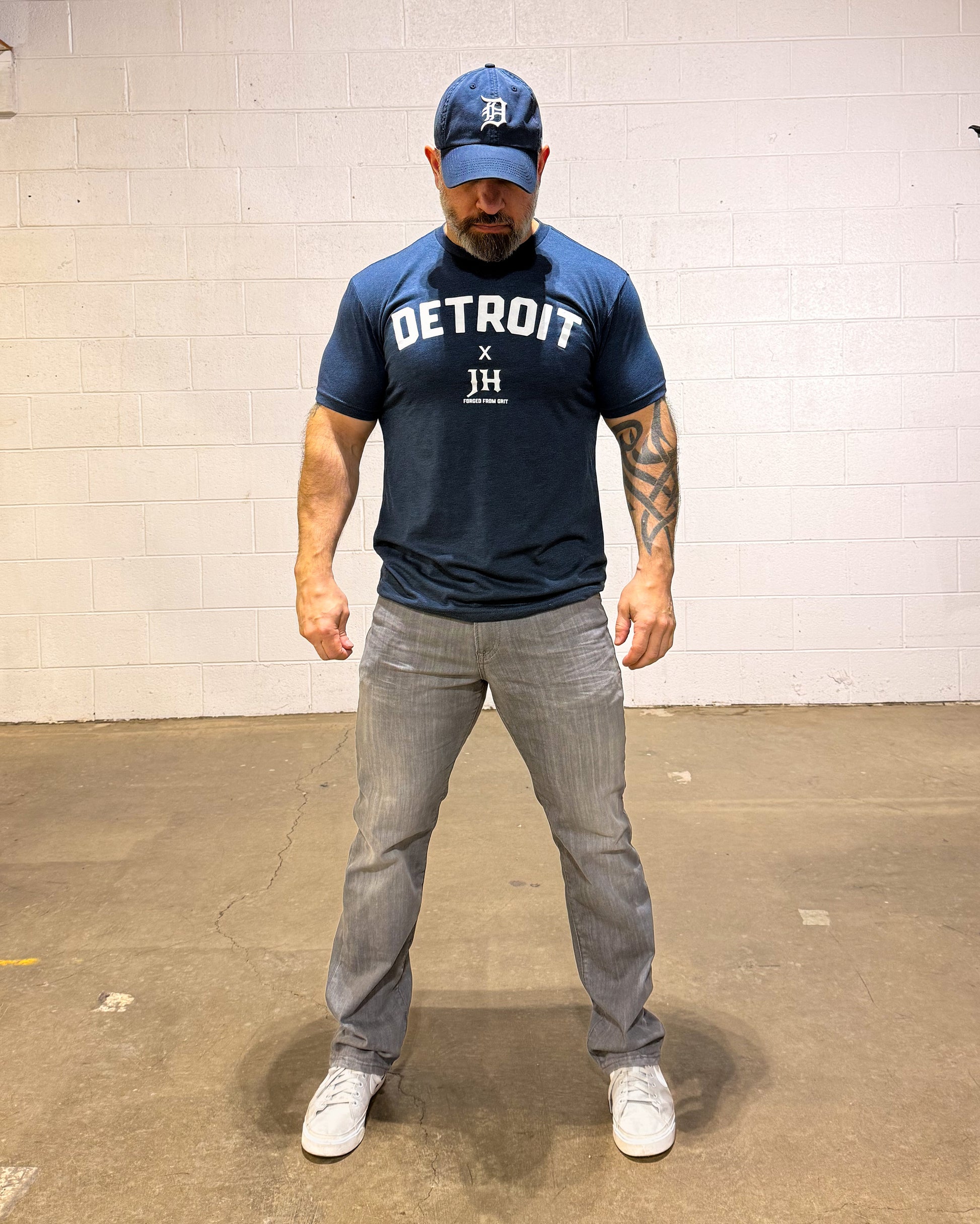Man wearing a navy blue 'Detroit' t-shirt and gray pants standing against a white brick wall.