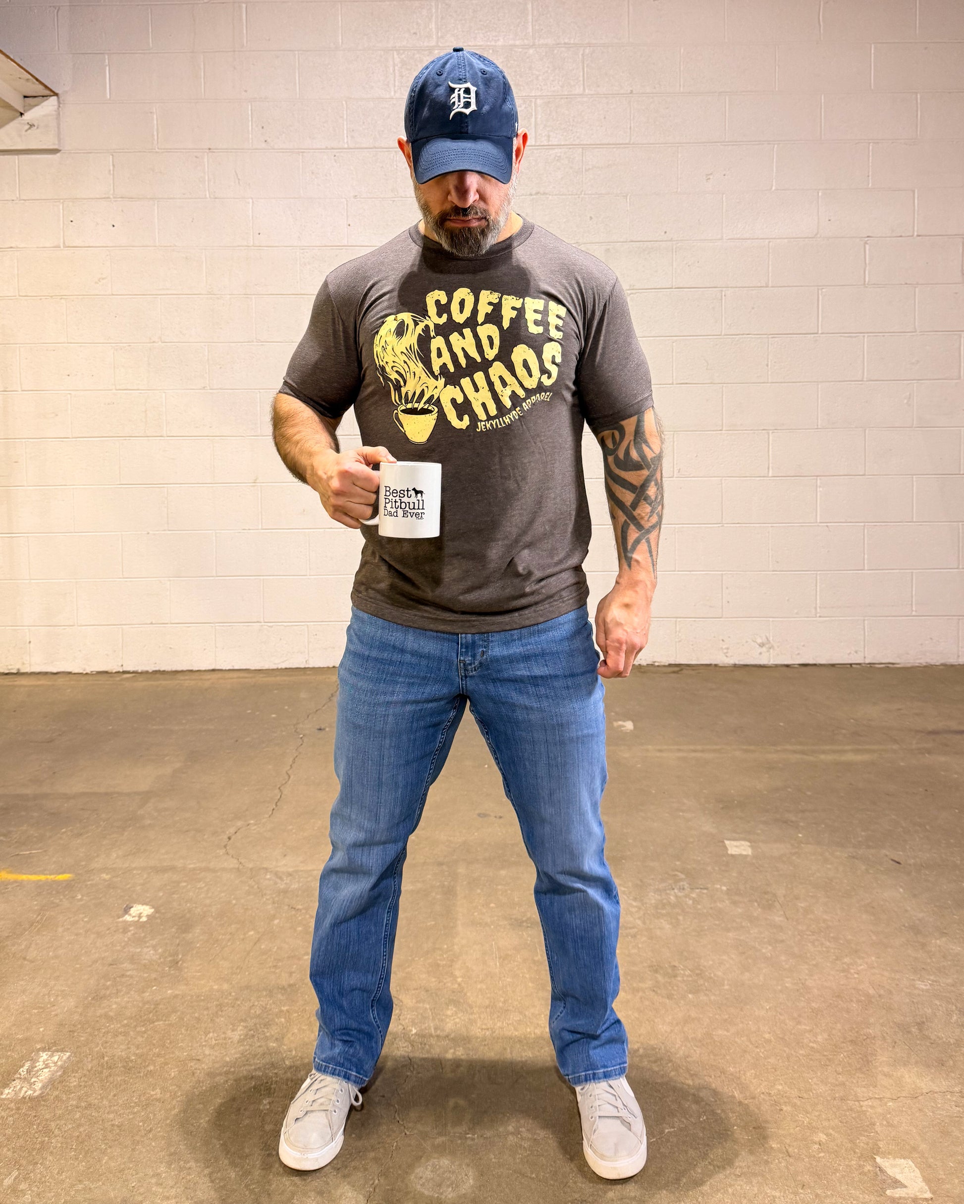 Man wearing a gray t-shirt with 'Coffee and Chaos' text, blue jeans, and a blue cap holding a coffee mug.