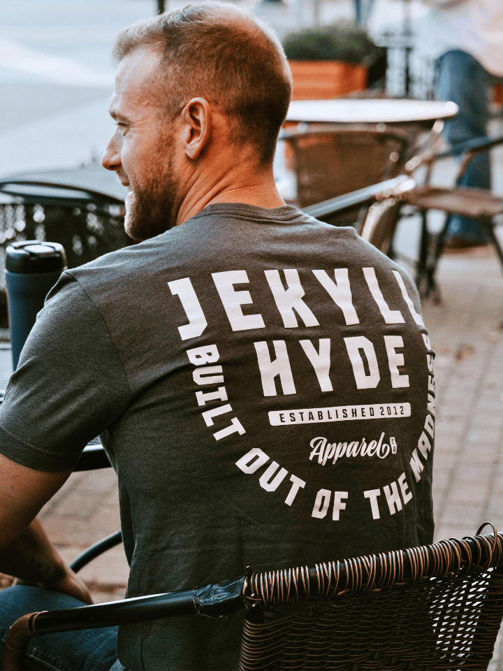 Man sitting at a table with his back to the camera wearing a grey shirt with Jekyllhyde in white print. 