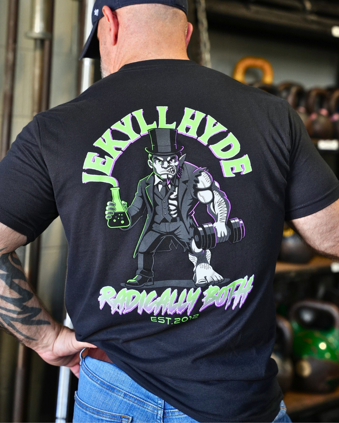 A man viewed from behind wearing a black graphic t-shirt with a large Jekyll Hyde design in green, gray, and white, set against a gym background with kettlebells.