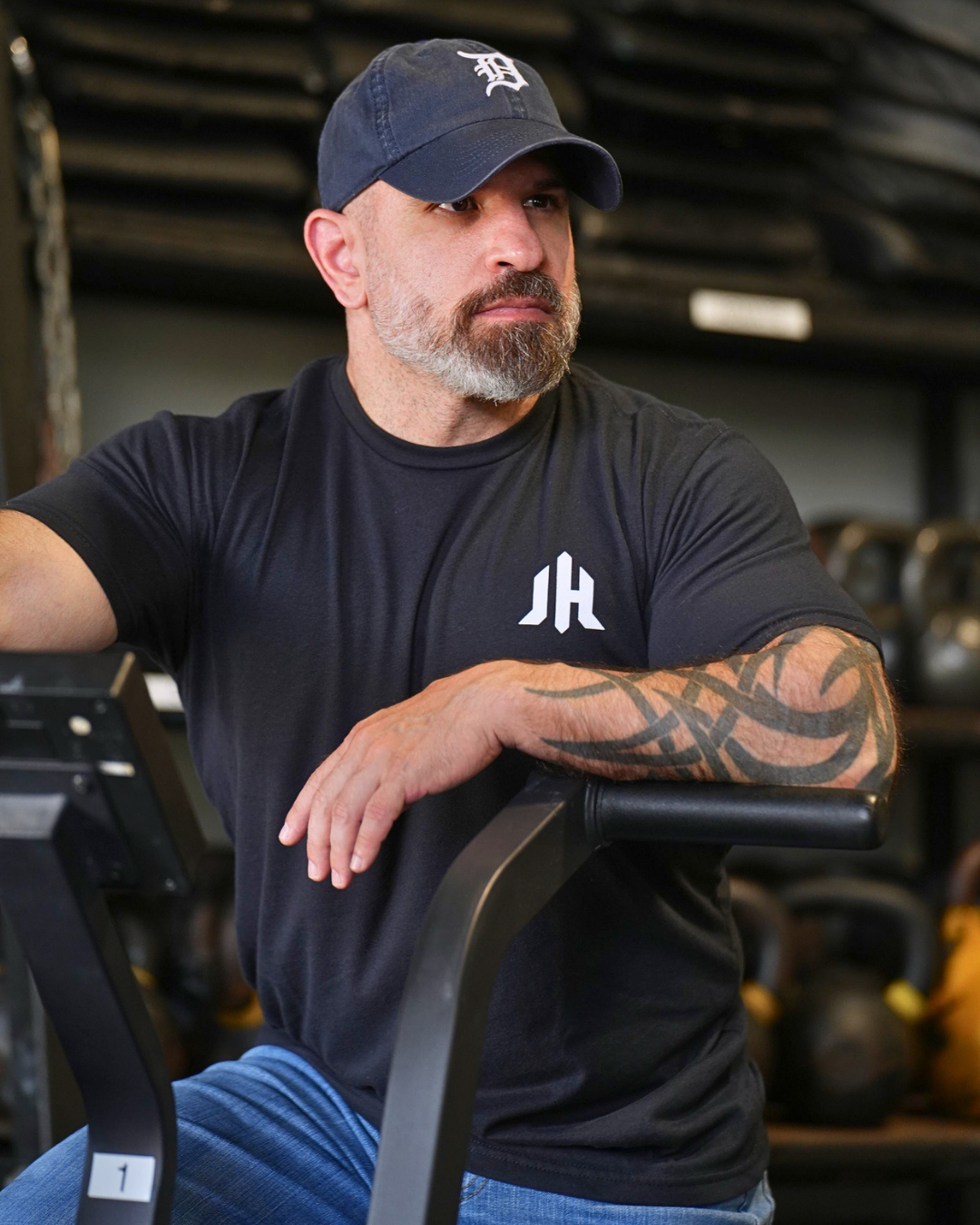 A man in a gym wearing a black t-shirt with a white stylized 'JH' logo on the chest, a navy blue baseball cap, and blue jeans, leaning on a treadmill.