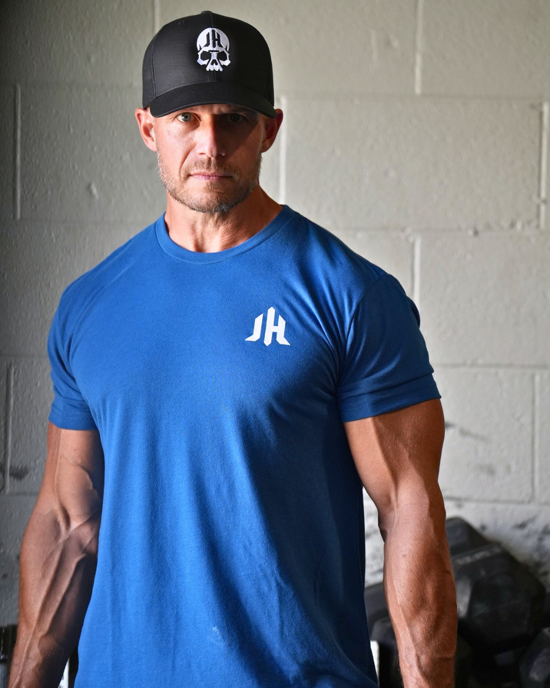 A muscular man wearing a bright blue short-sleeve athletic t-shirt with a white stylized 'JH' logo on the left chest, paired with a black baseball cap featuring a white skull graphic. He is standing against a light gray concrete block wall.