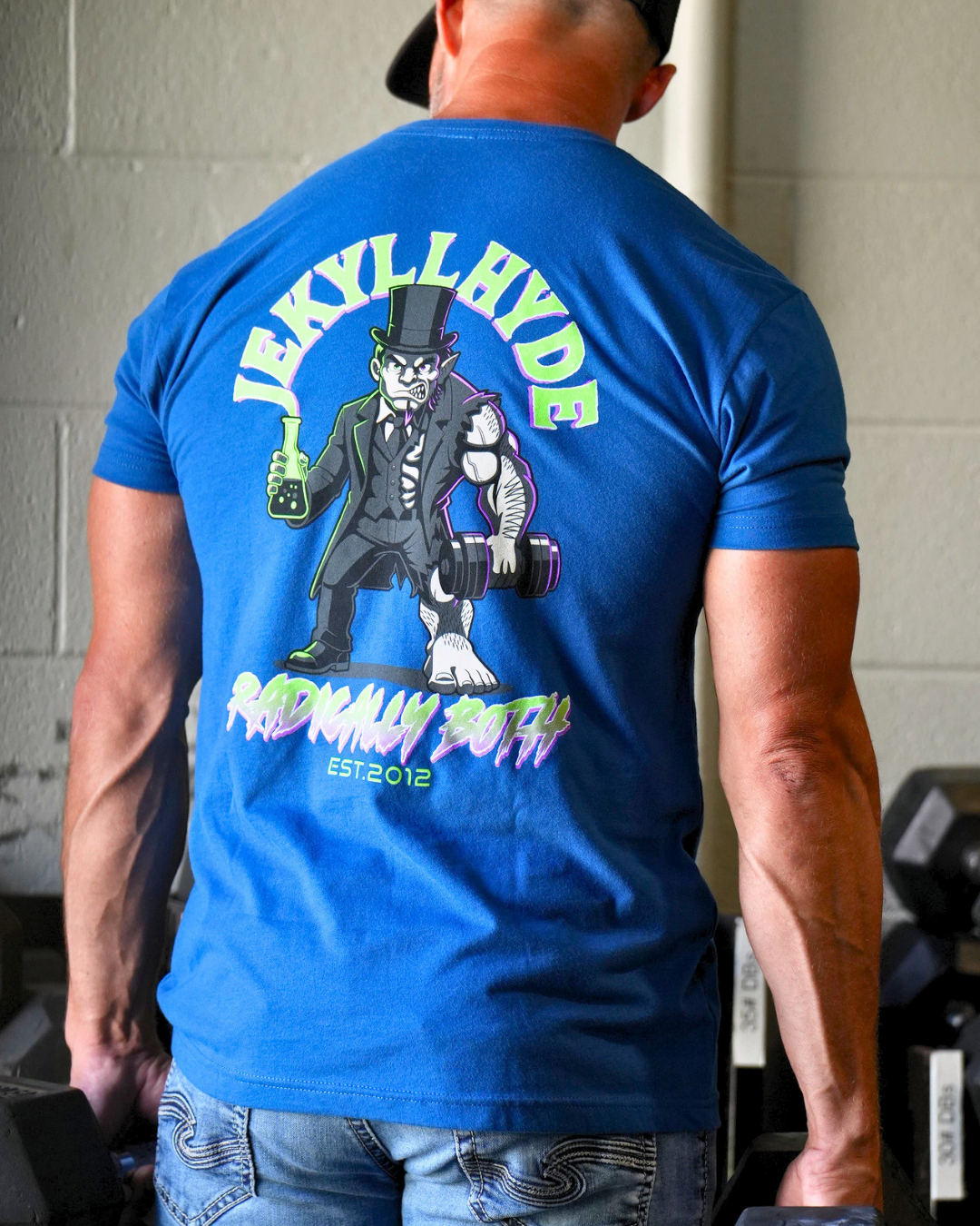 A man viewed from behind wearing a bright blue t-shirt with a large graphic print on the back. The graphic features the text 'JEKYLL HYDE' and 'RADICALLY BOTH EST 2012' surrounding an illustration of a skeleton in a top hat holding a flask and a barbell.