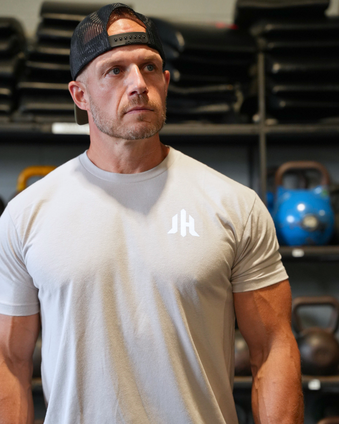 A muscular man in a gym wearing a light gray crewneck t-shirt with a white stylized 'JH' logo on the chest, paired with a black mesh-back baseball cap.