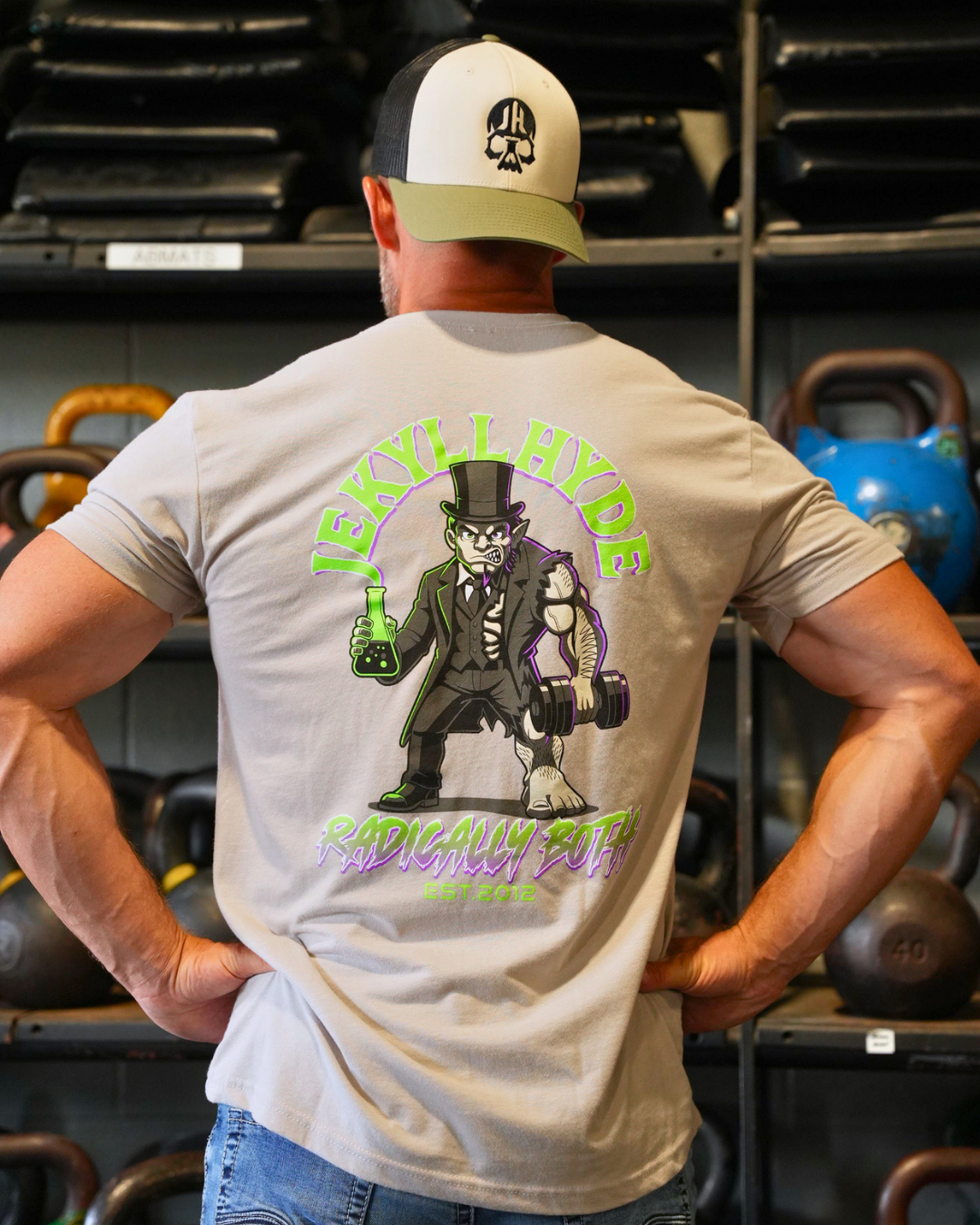 A man viewed from behind wearing a light gray t-shirt with a large graphic print of a cartoon character and the text 'JEKYLL HYDE RADICALLY BOTH' in green and purple, set in a gym with kettlebells on the background.