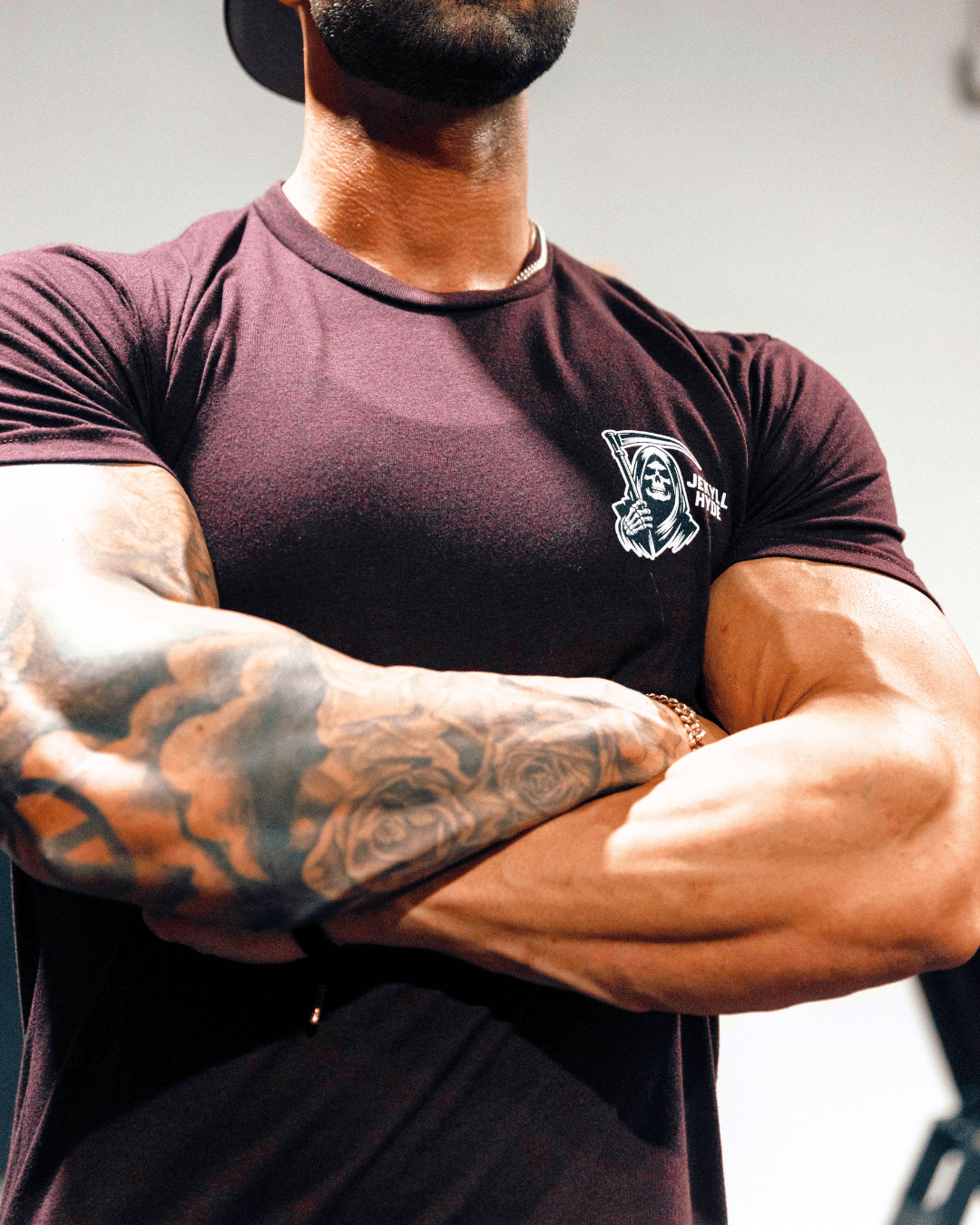 Person wearing a dark maroon t-shirt with a logo, arms crossed, on a plain background