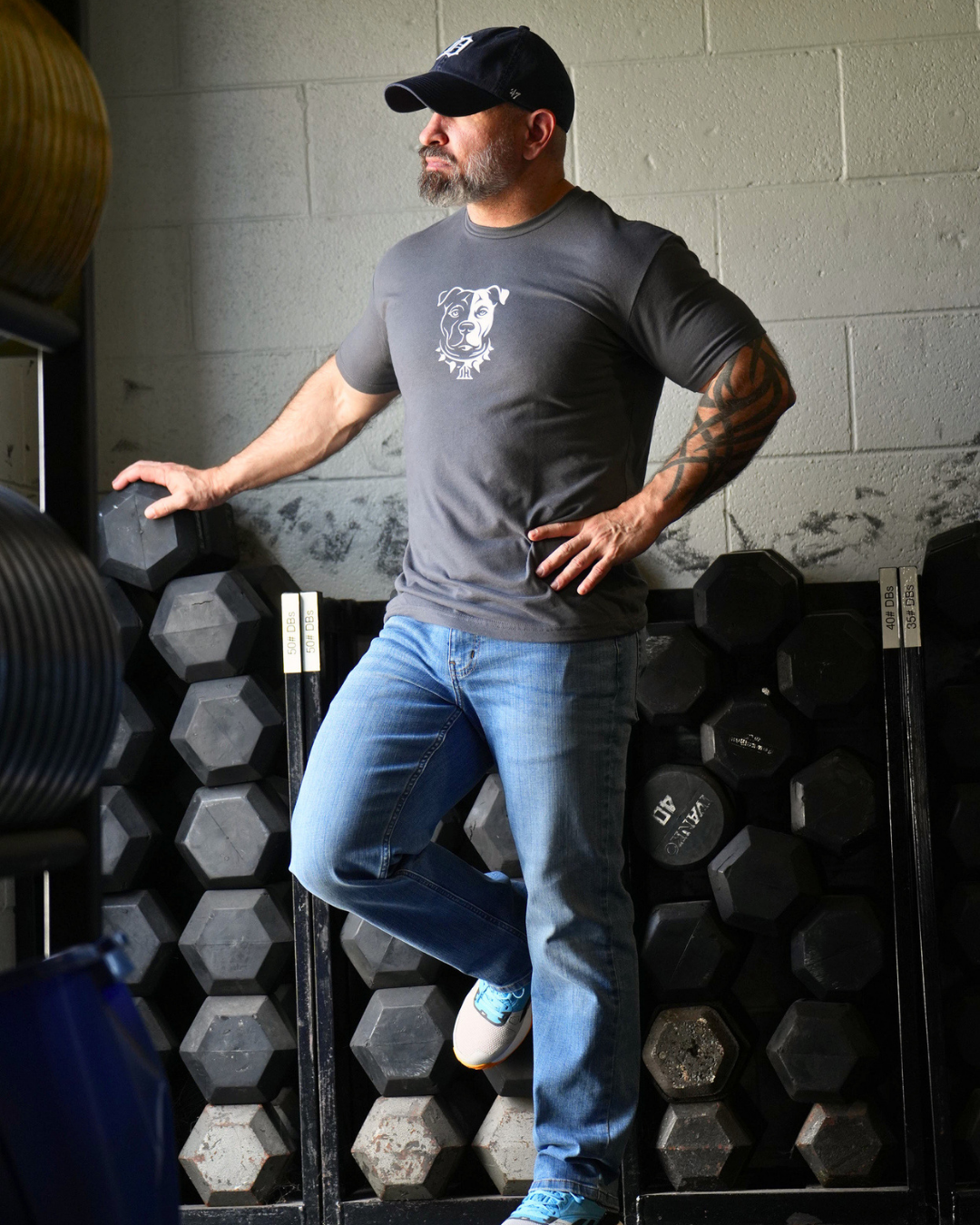 A man in a gym wearing a dark gray t-shirt with a white dog graphic, blue jeans, and a black cap, posing in front of a rack of hex dumbbells.
