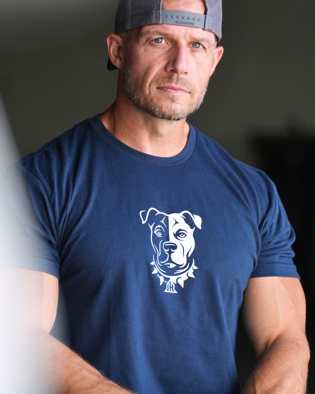 A muscular man wearing a navy blue t-shirt with a white graphic of a pitbull dog wearing a collar, paired with a gray baseball cap, in a dimly lit indoor setting.