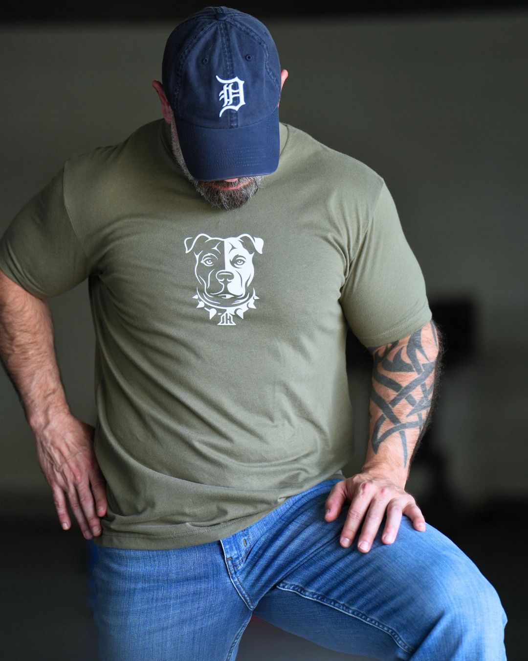 A man wearing an olive green t-shirt with a white pitbull graphic on the chest, blue jeans, and a navy blue baseball cap with a white 'D' logo, posing in a dimly lit indoor setting.
