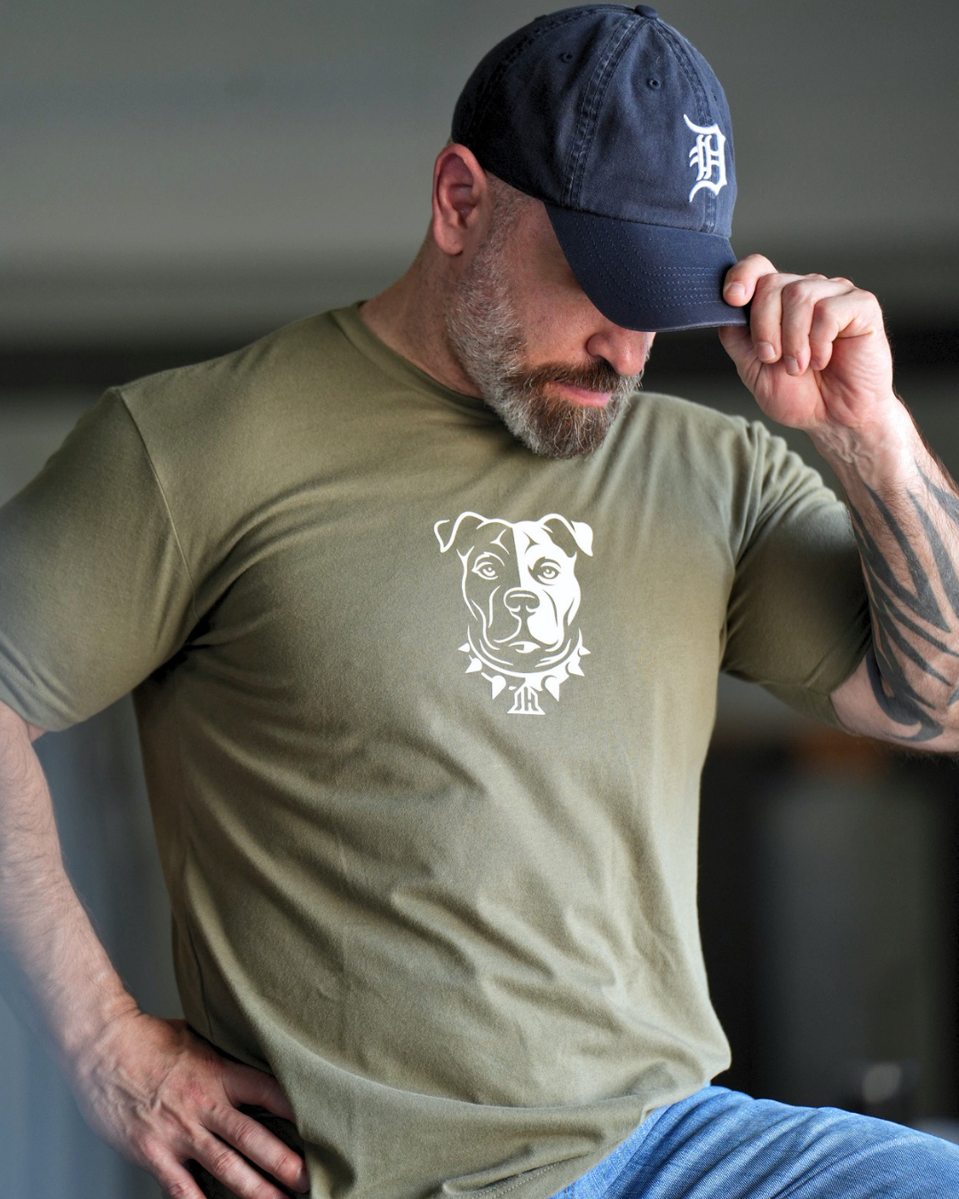A man touching his baseball hat wearing an OD green shirt with a Pitbull graphic on it. 