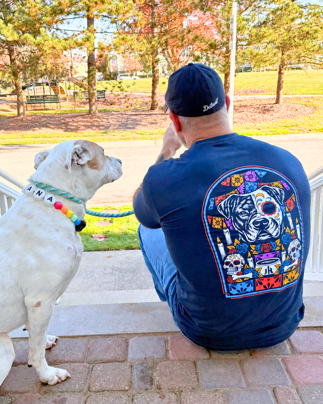 Muscular man sitting with a dog with a navy shirt with a pitbull ofrenda on the back. 