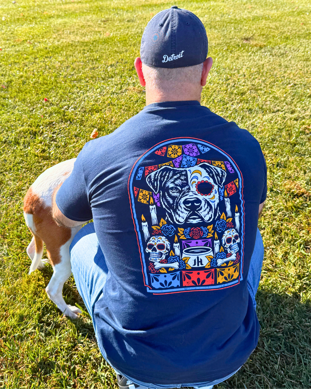 Muscular man sitting with a dog with a navy shirt with a pitbull ofrenda on the back. 