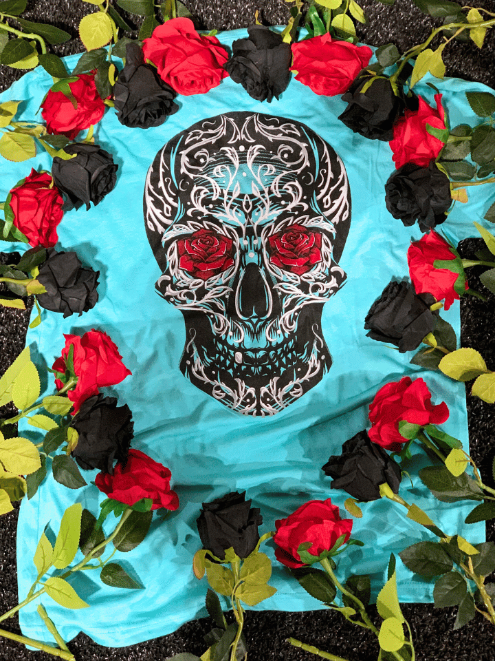 A blue shirt with a sugar skull on it laying on the ground with black and red roses surrounding it. 